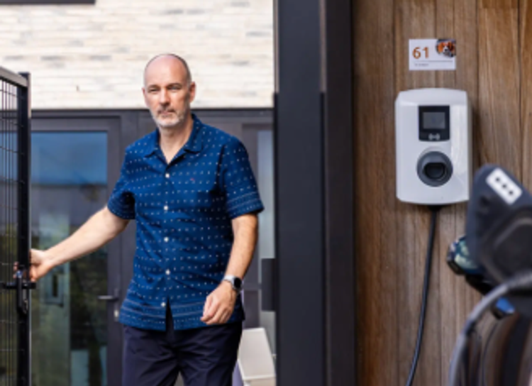 Man met blauw hemdje staat voor zijn deur bij oplaadpunt aan een houten wand en e-wagen.