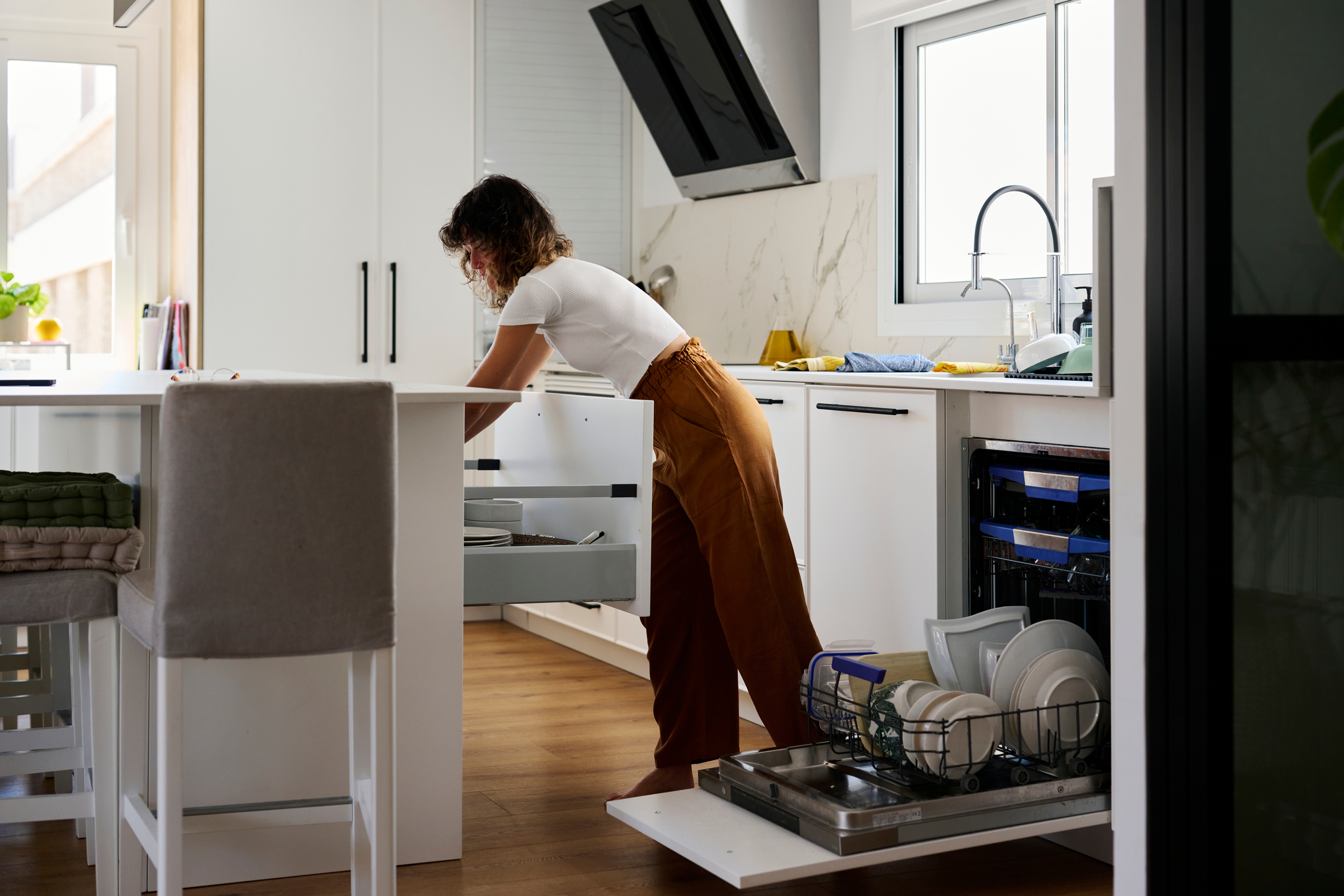 Moderne keuken, de vaatwasser met schone vaat staat open, dame met bruine brede broek en witte top laadt deze uit en zet het servies weg in de kast. Dame laadt de schone vaat