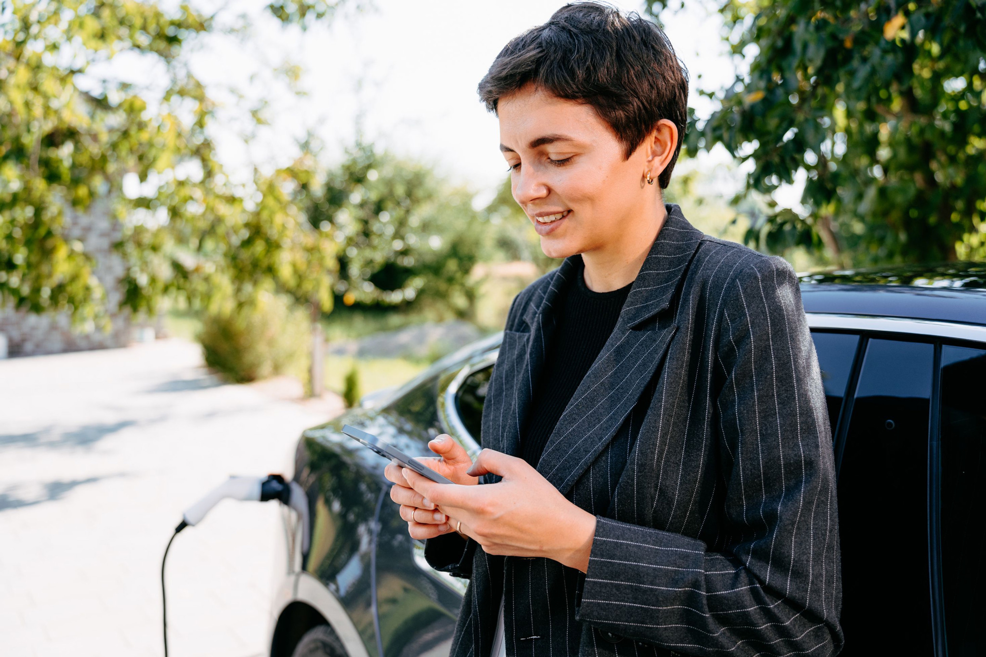 Dame staat bij haar e-wagen die oplaadt, ze kijkt op haar phone.