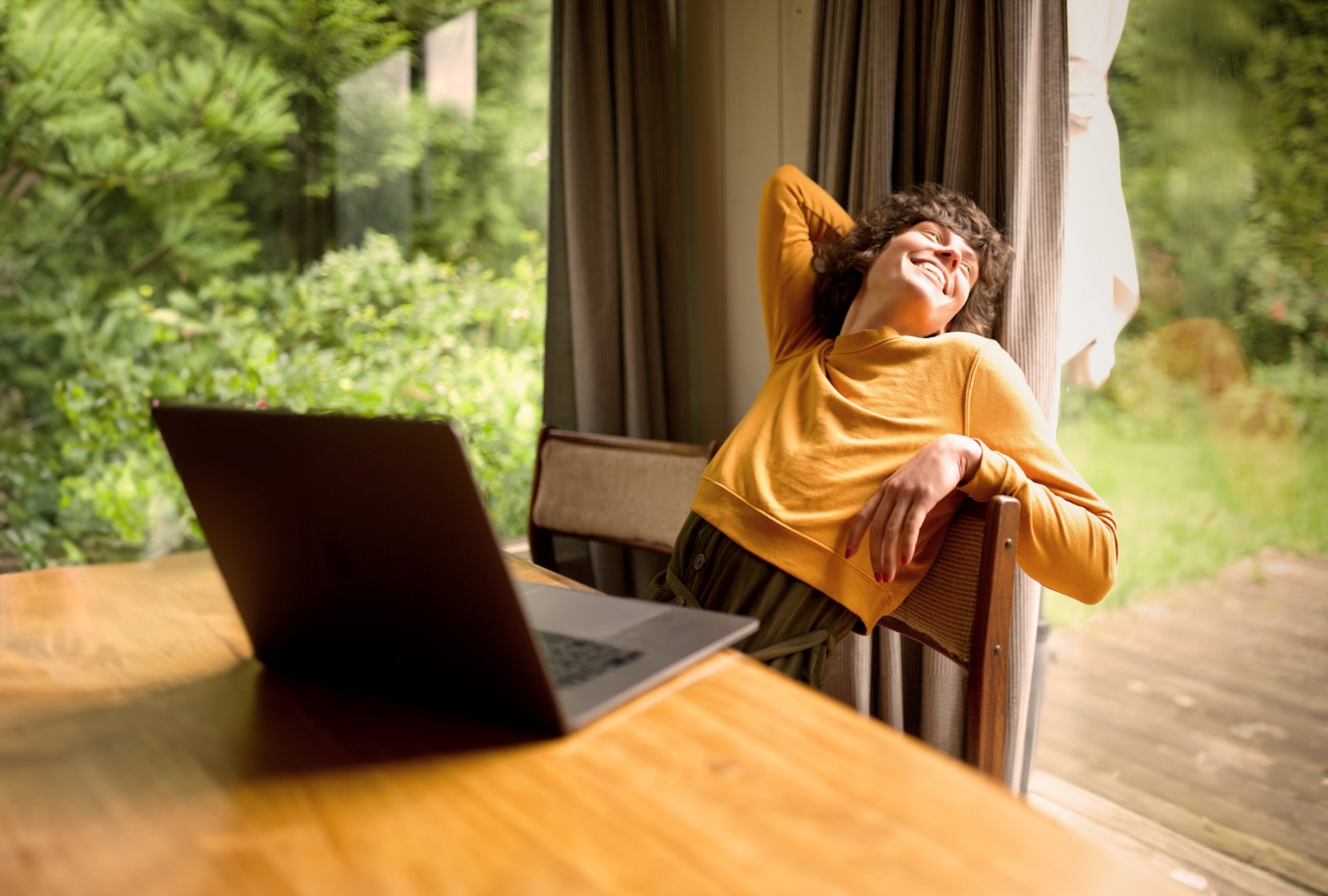Jonge vrouw met oranje trui aan, laptop staat open op tafel, zij leunt ontspannen en lachend achterover.