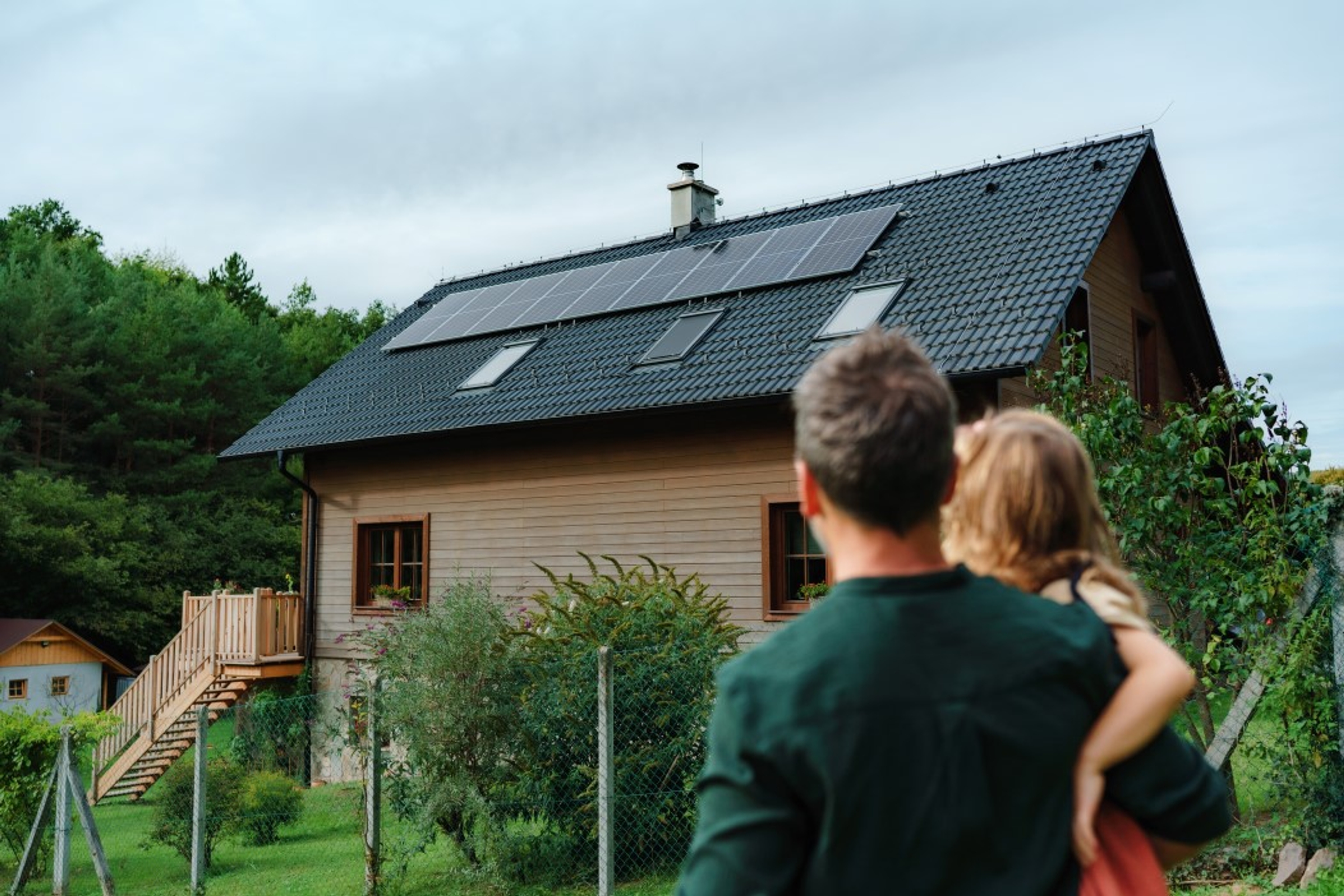 Un homme portant un enfant regarde une maison en bois équipée de panneaux solaires sur le toit, entourée de verdure