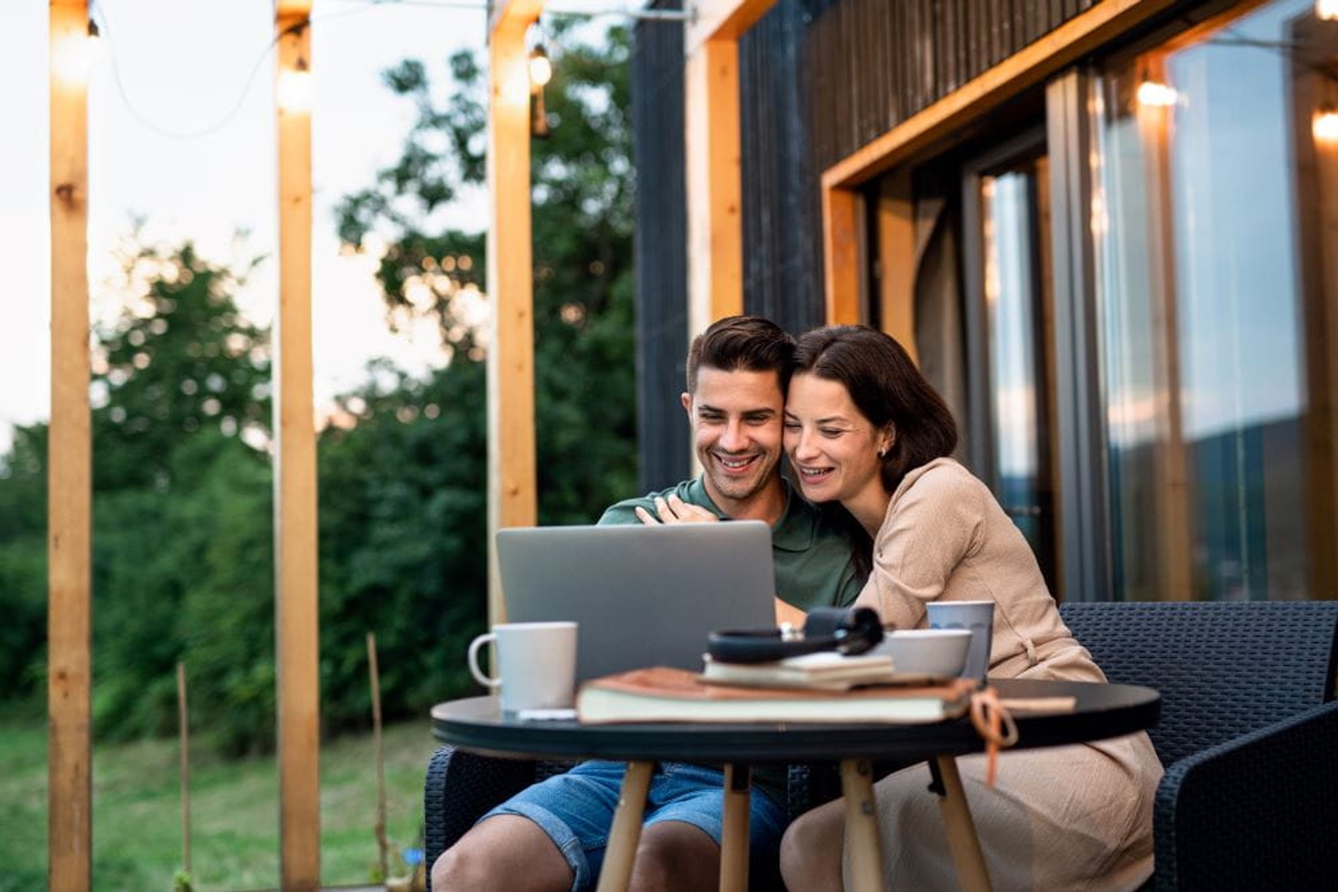 Koppel zit buiten aan hun houten huis, kijkt lachend op de laptop, houten balken en lampjes in de achtergond.