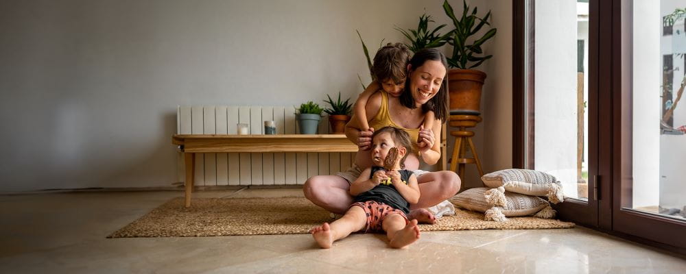 Jonge vrouw en 2 kindjes in de living op de grond, een van de kindjes eet een frisco.