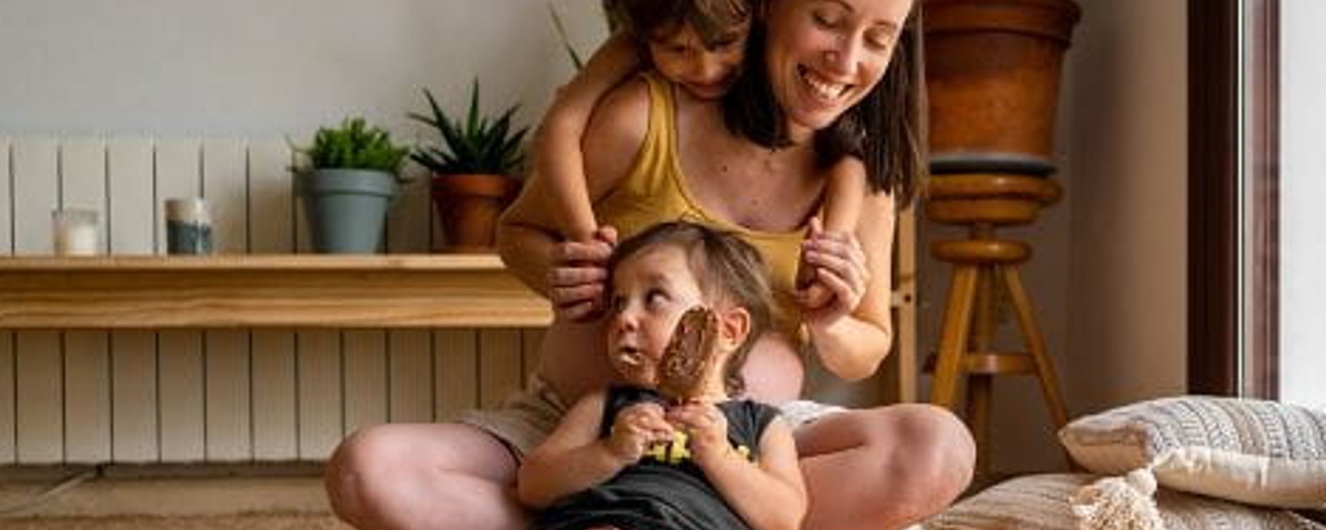Jonge vrouw en 2 kindjes in de living op de grond, een van de kindjes eet een frisco.