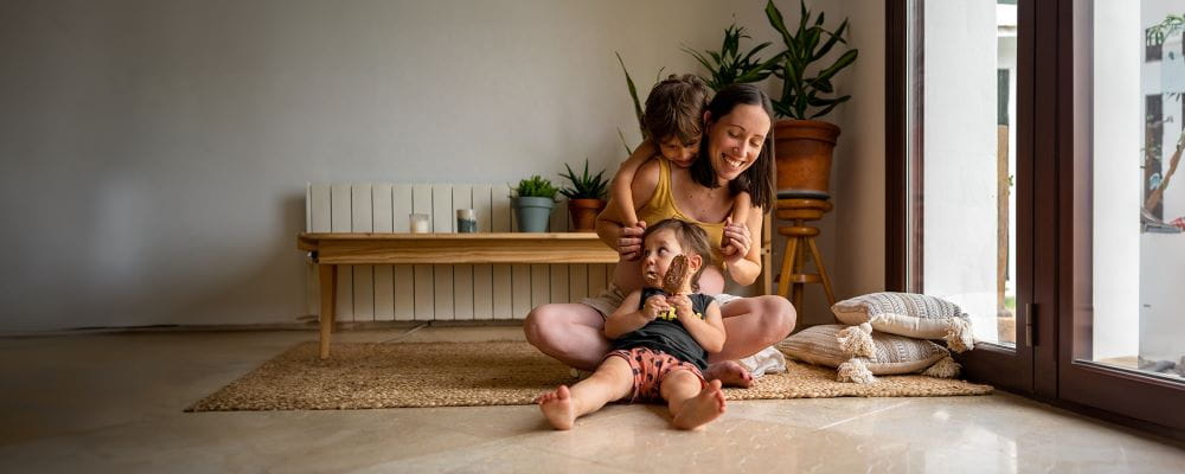 Jonge mama speelt met 2 kindjes in de woonkamer op het tapijt, verwarming op de achtergrond.