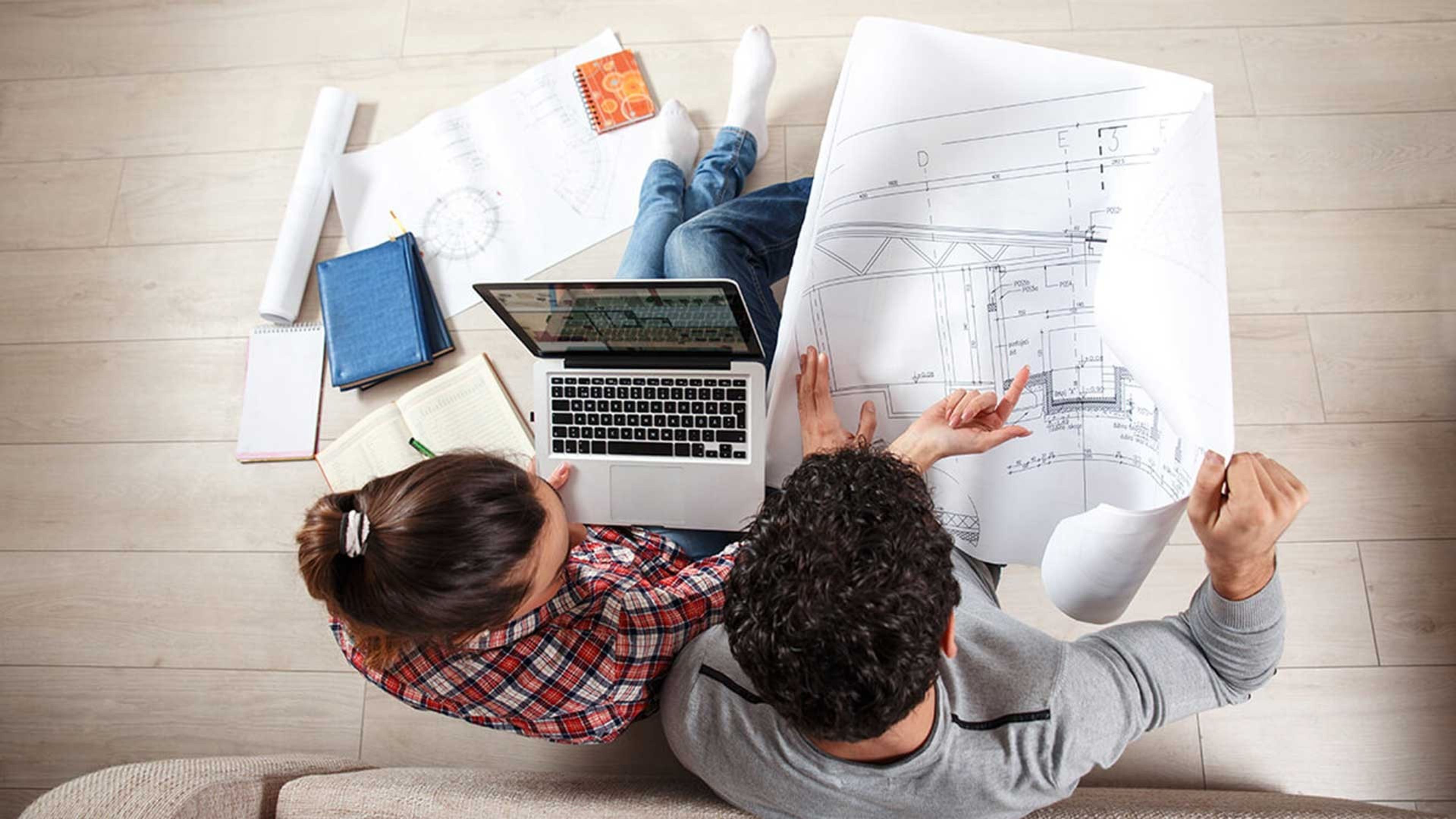 Koppel van boven gezien zit op de grond in hun living met architectenplan en laptop.