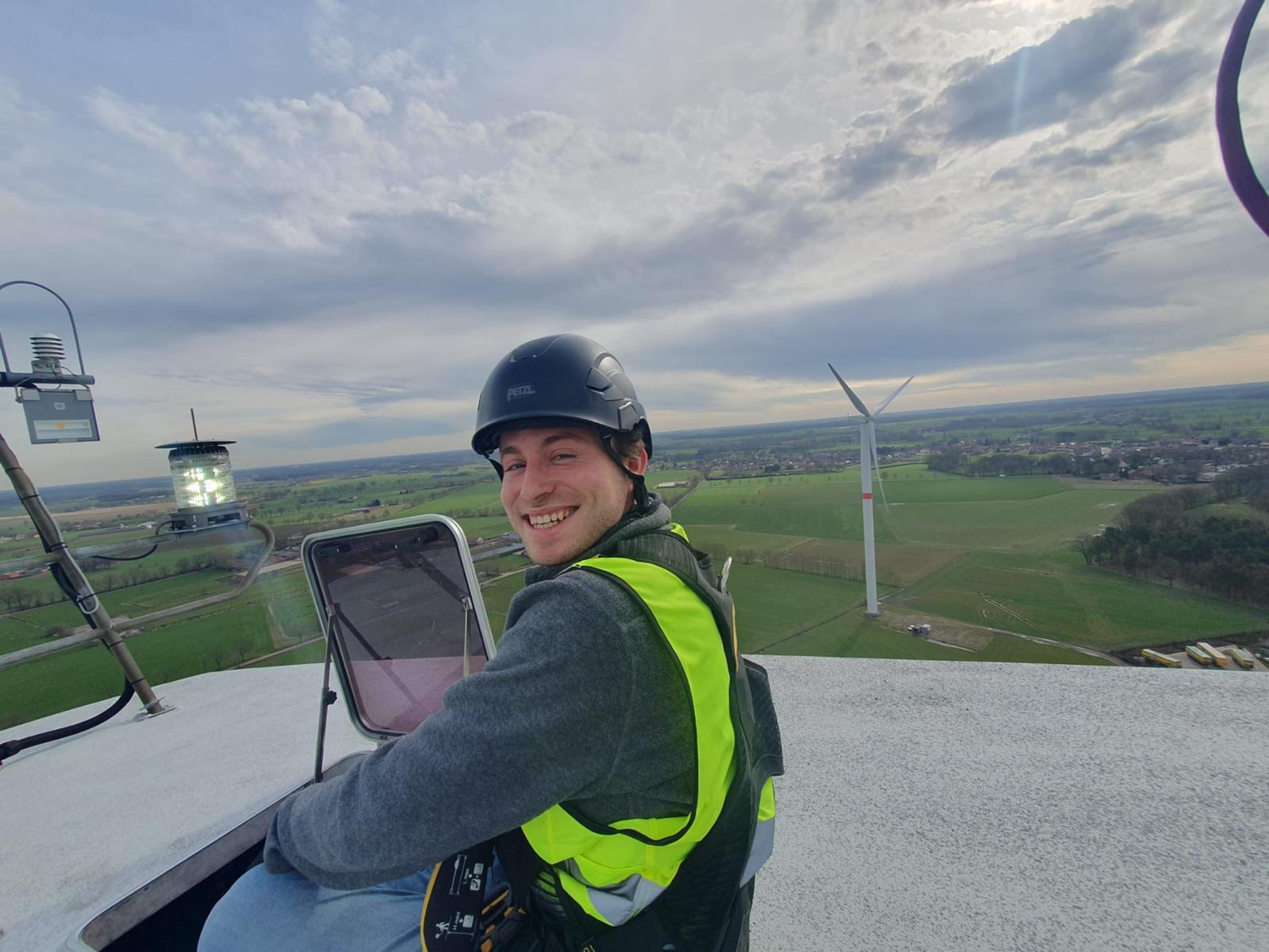 Een persoon in een veiligheidsvest en helm glimlacht terwijl hij bovenop een windturbine zit. Achter hem strekt zich onder een bewolkte hemel een uitgestrekt landschap uit met velden en nog meer windturbines.