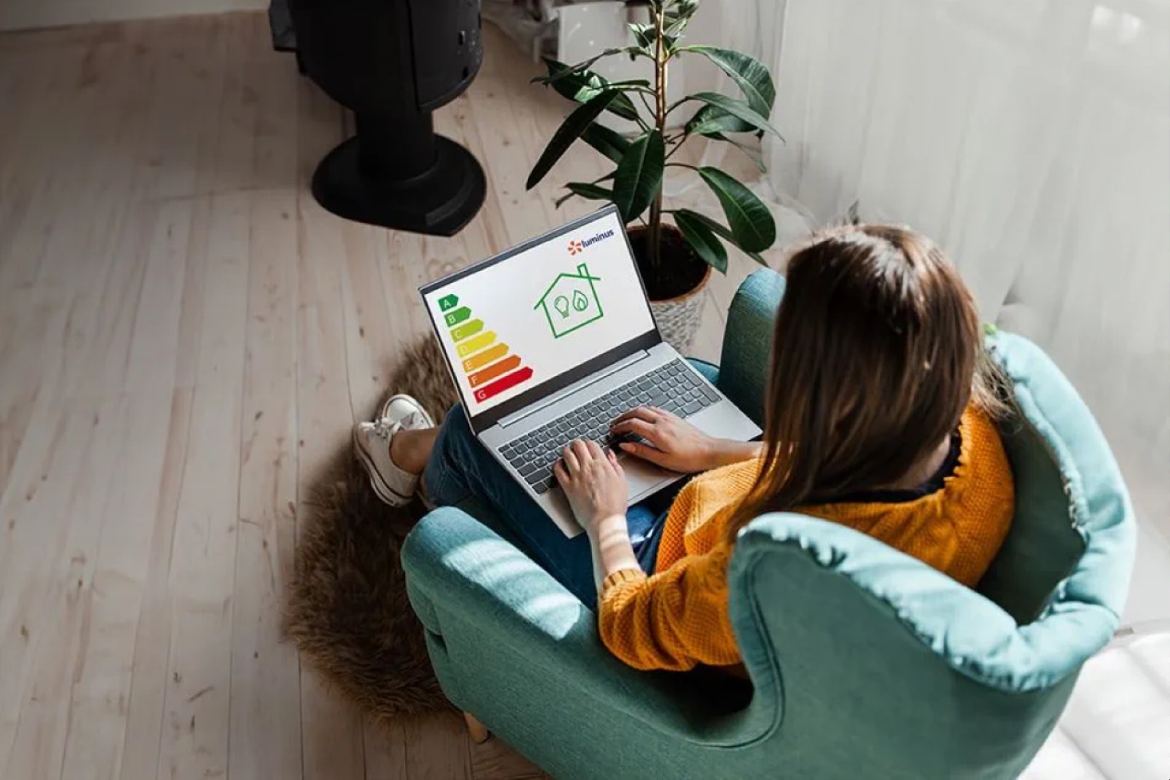 Dame zit in lichtgroen zeteltje en kijkt op laptop, in het scherm de lijntekening van een huisje en de gekleurde balken van energielevels A, B, C ..., alles van bovenaf gezien. 