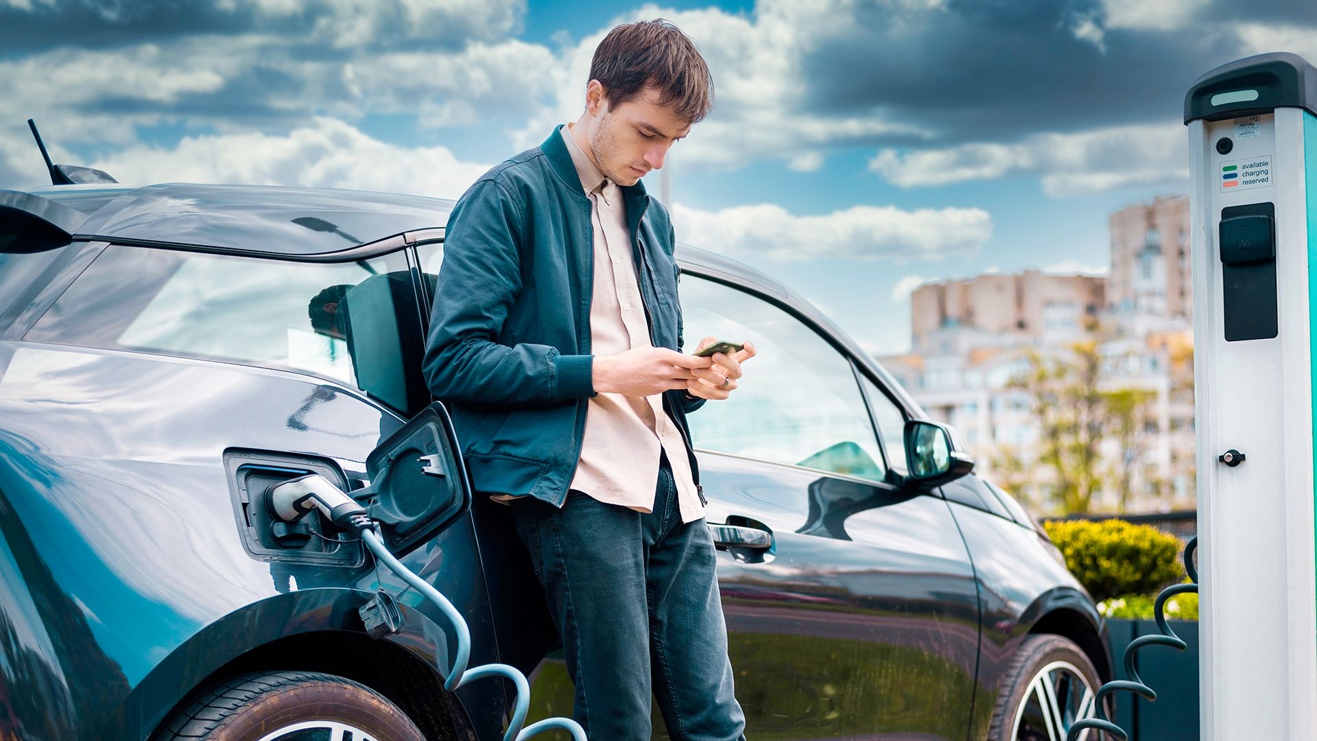 Jongeman kijkt op zijn gsm en staat bij elektrische wagen die oplaadt.
