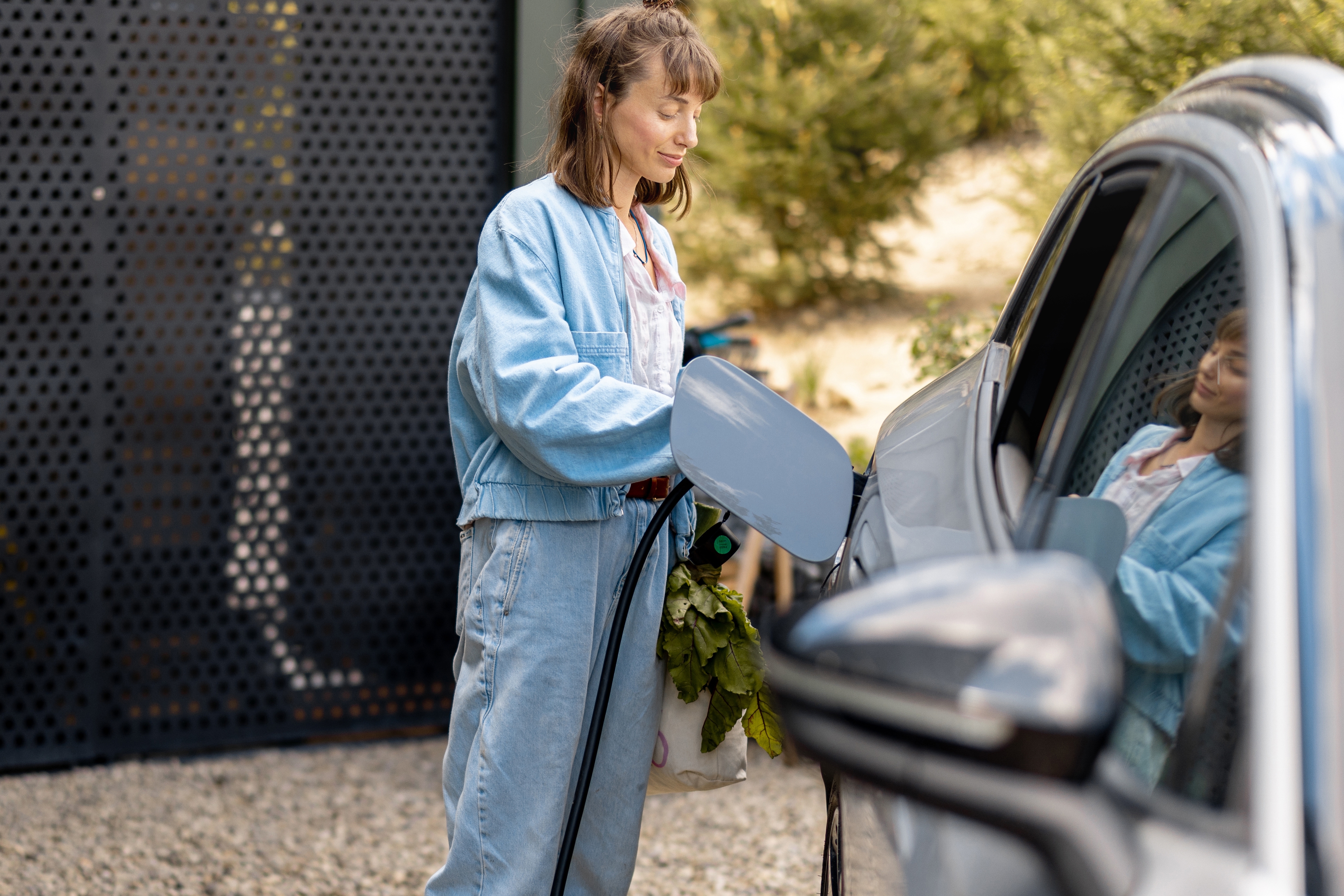 Jongedame laadt haar e-wagen thuis op, zwarte metalen geperforeerde wand en boompjes in de yuin.