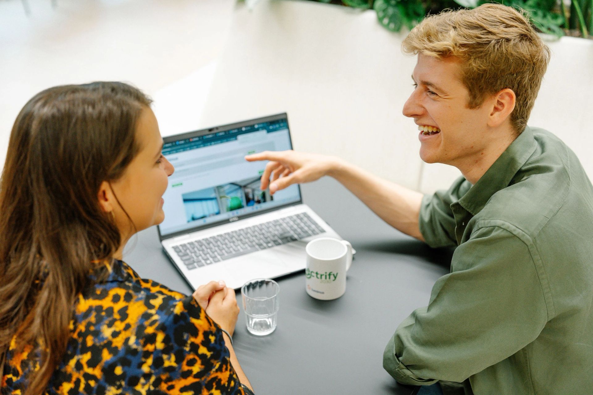 Een man en een vrouw zitten aan een tafel, glimlachen en bespreken iets op een laptopscherm. Op het bureau staan een mok en een glas. De sfeer straalt een positieve samenwerking uit.