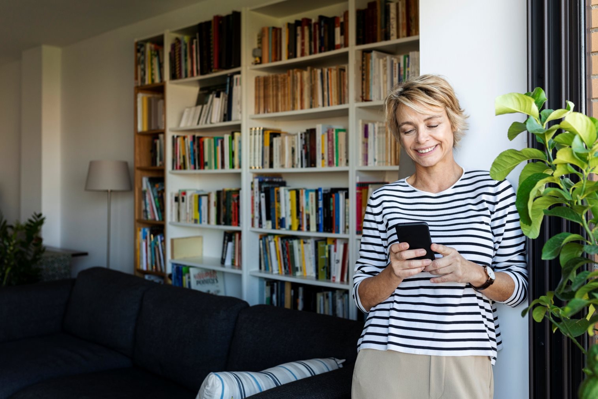 Femme souriante debout dans un salon moderne, tenant un smartphone devant une bibliothèque remplie de livres