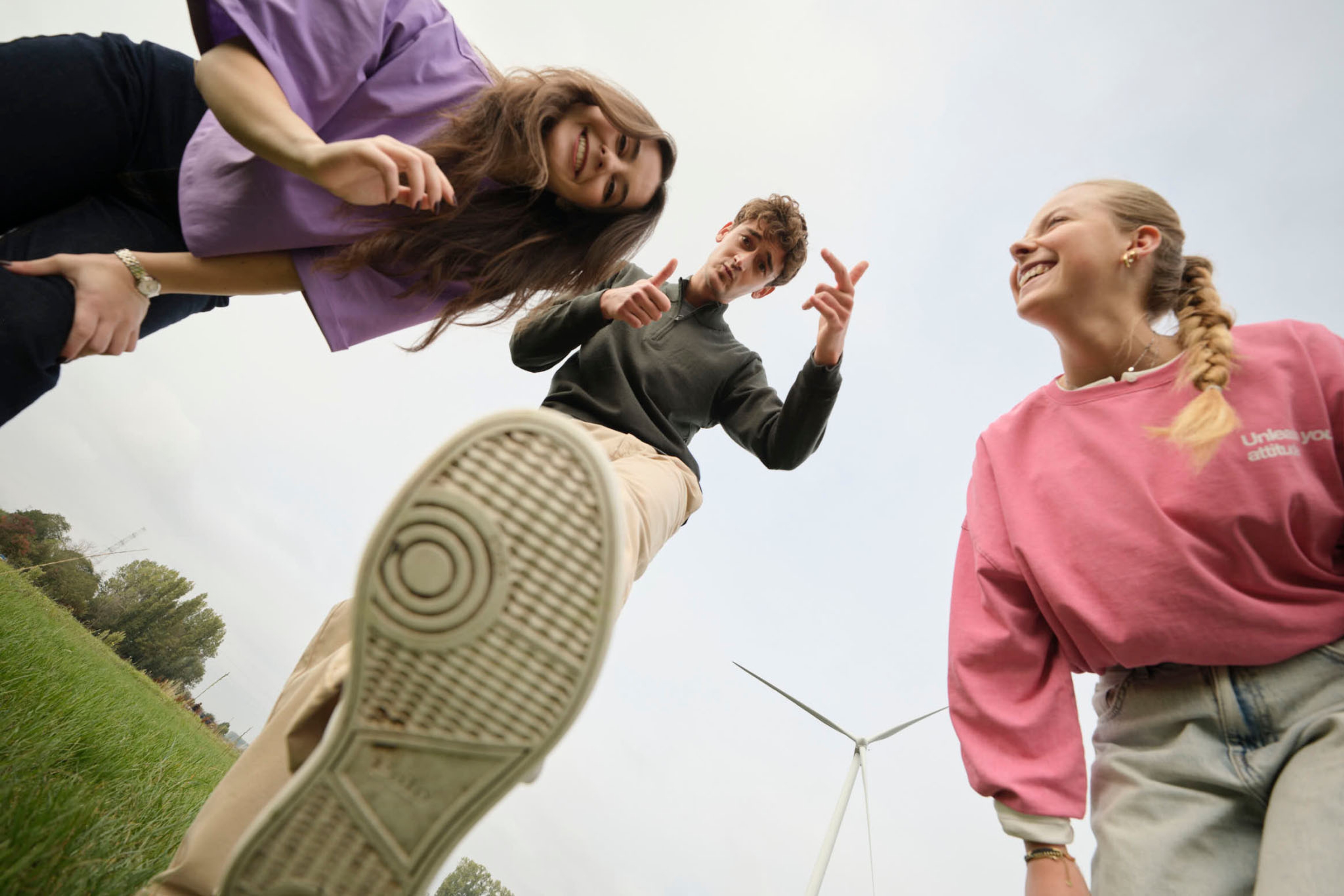 Een grote, diverse groep mensen in kleurrijke shirts glimlachen en zwaaien enthousiast voor een bord met het opschrift ‘GENERATION’ in een lichte, industriële ruimte.