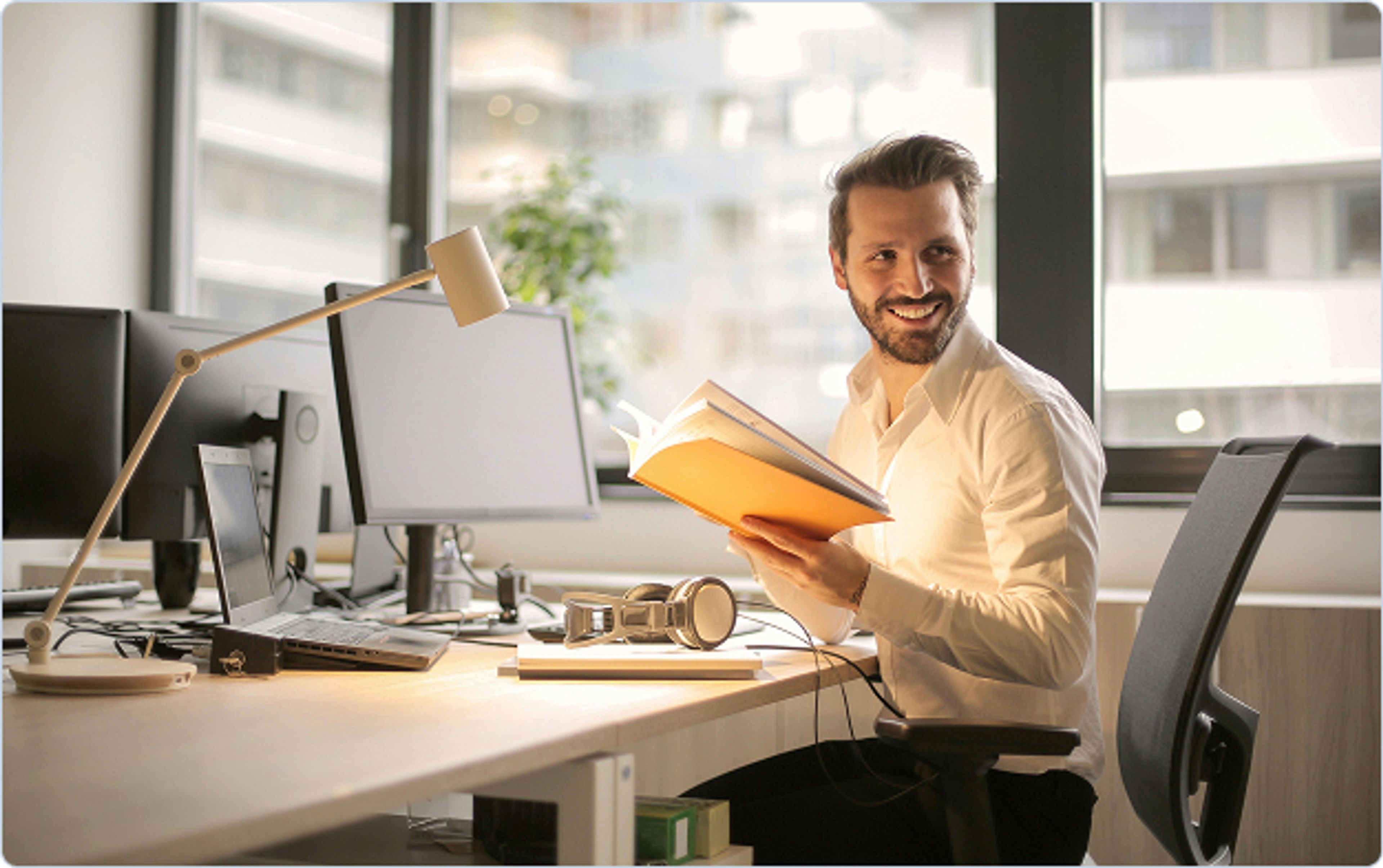 Homme souriant assis à un bureau moderne, tenant un livre ouvert à la main, avec plusieurs écrans d’ordinateur et une lampe de bureau, dans un bureau lumineux avec de grandes fenêtres