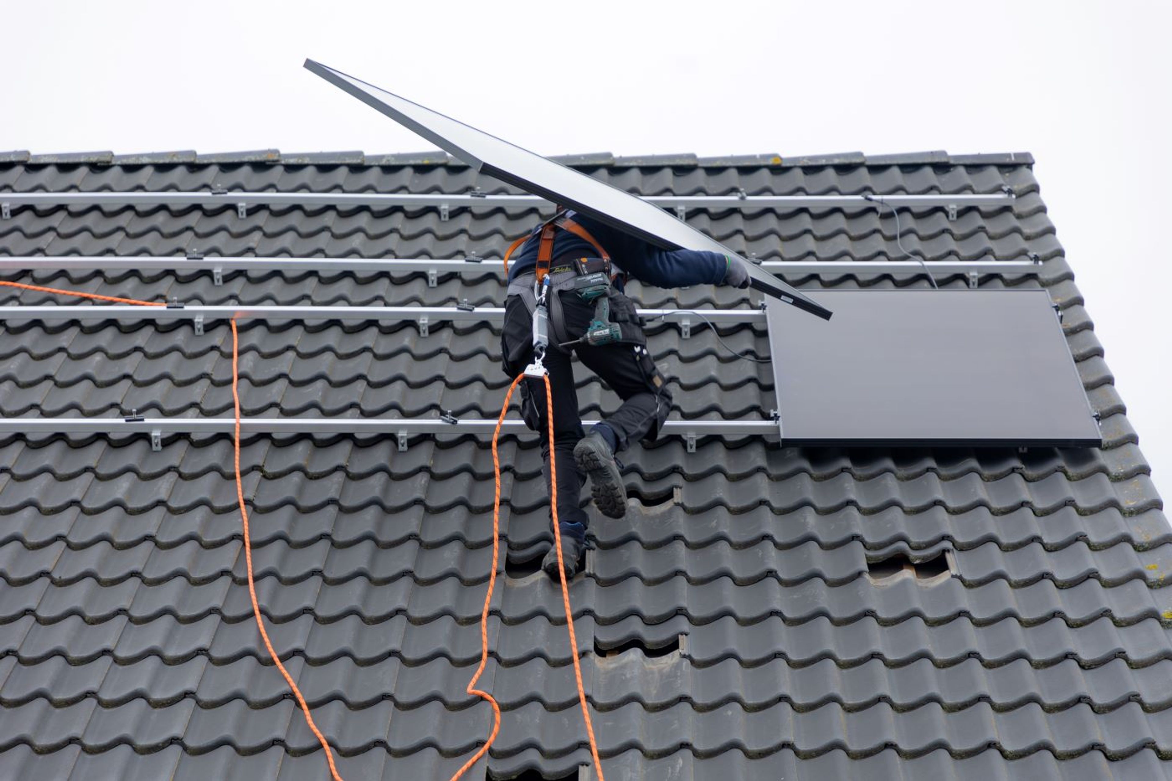 Beveiligde technieker klimt op dak met zonnepaneel op de schouder, dat gaat hij zometeen installeren.