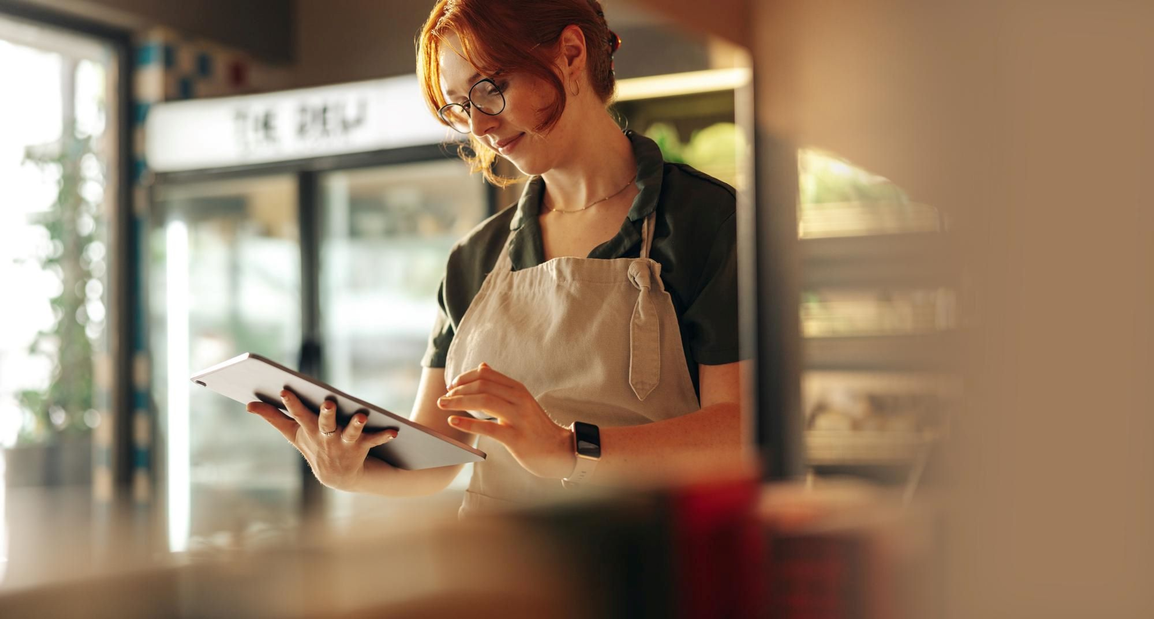 Dame met rood opgestoken haar en beige schort kijkt iets na op een tablet in een winkel met koelkasten.
