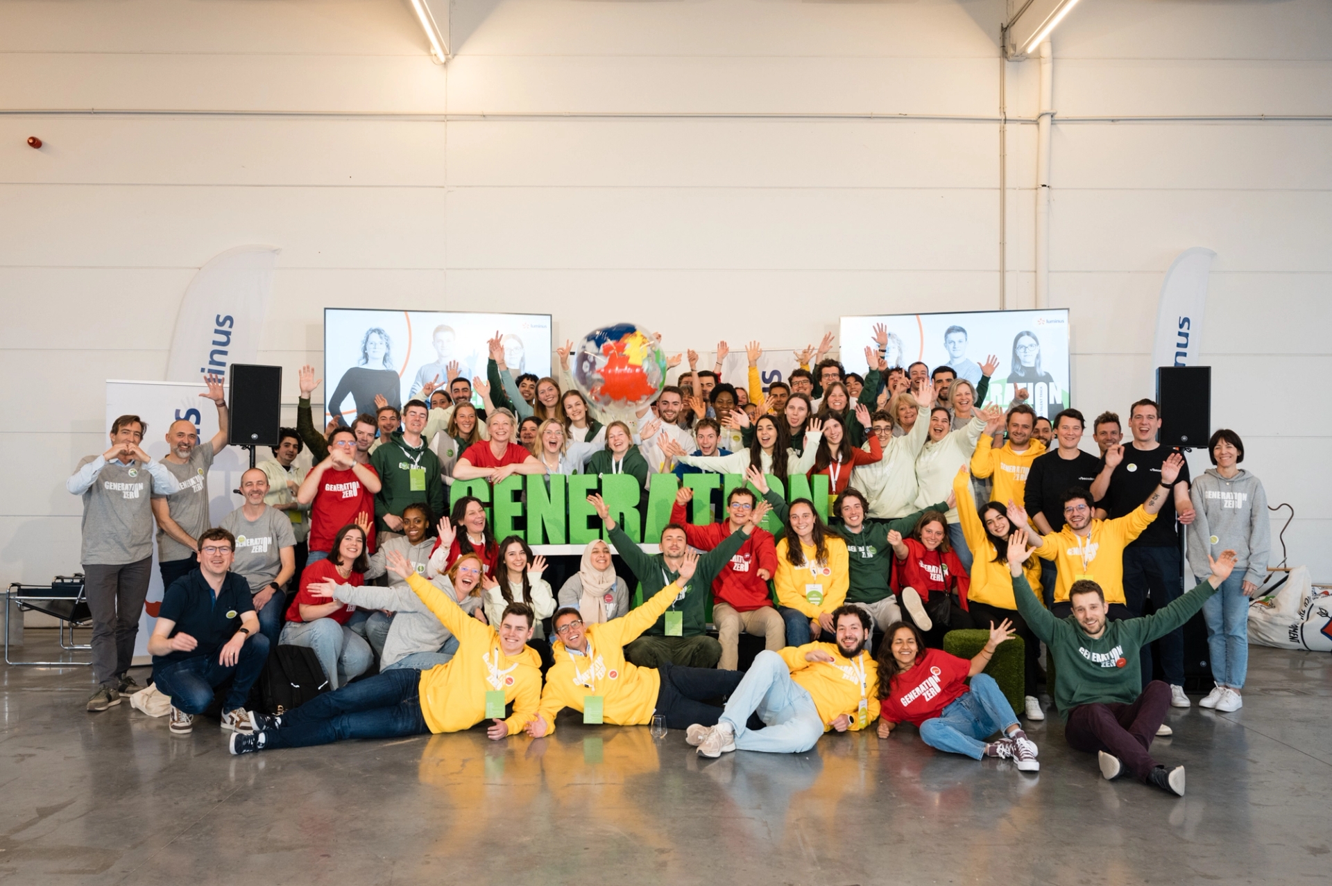 Un groupe important et diversifié de personnes vêtues de chemises colorées sourit et salue avec enthousiasme devant une enseigne « GENERATION » dans un espace industriel lumineux.