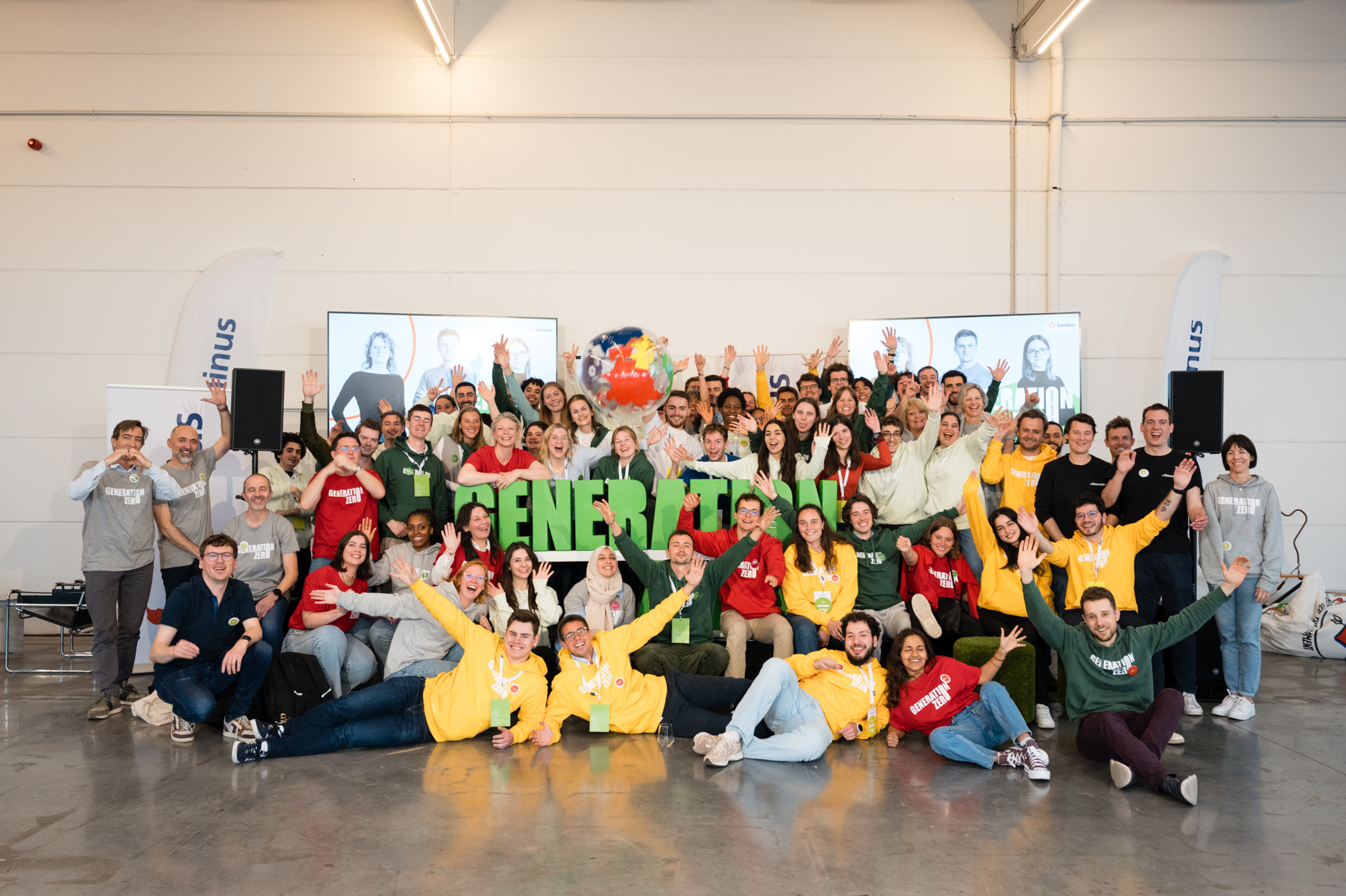 Un groupe important et diversifié de personnes vêtues de chemises colorées sourit et salue avec enthousiasme devant une enseigne « GENERATION » dans un espace industriel lumineux.