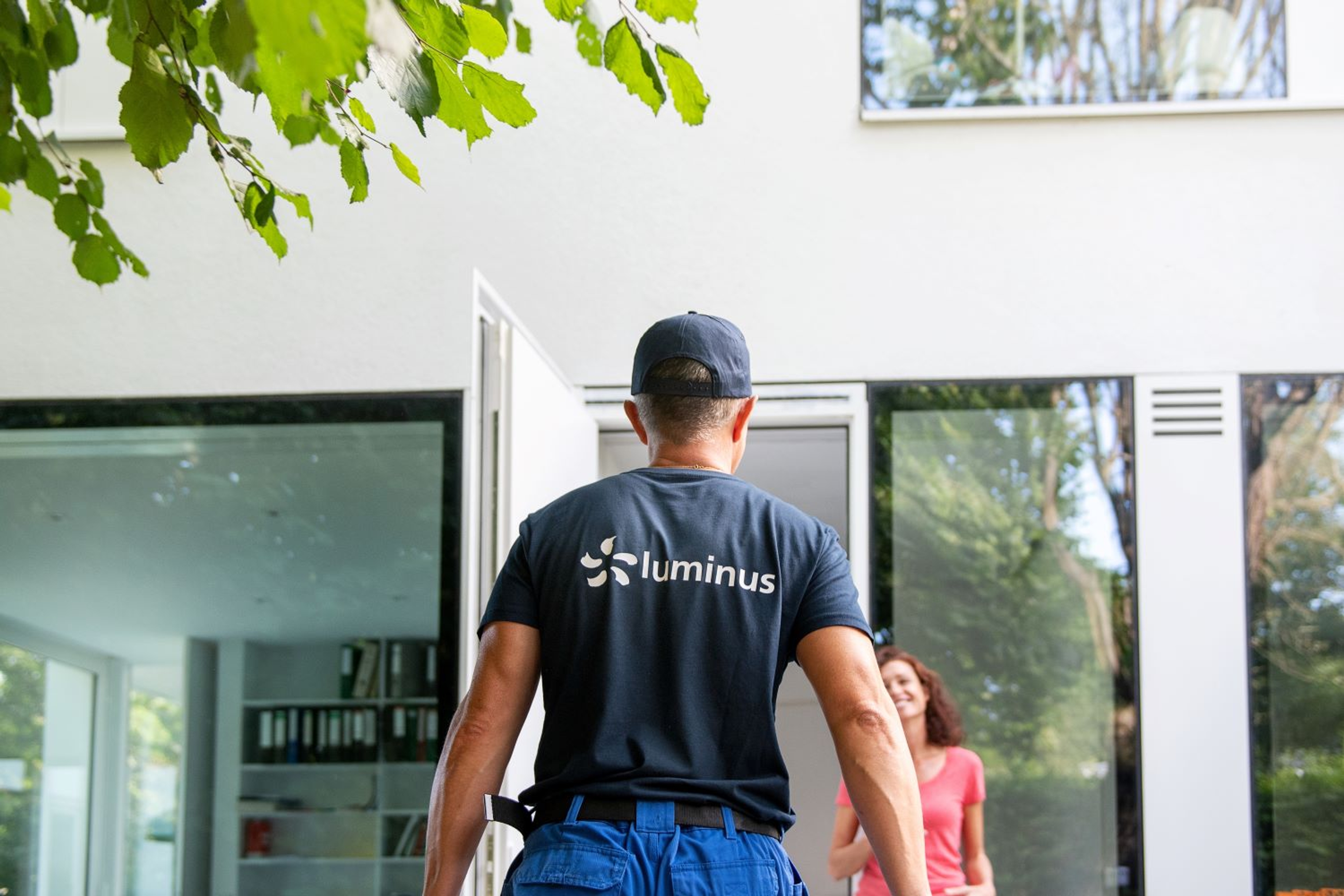 Technieker van Luminus komt aan bij wit modern huis. Dame met rode top wacht hem lachend op bij de open voordeur.