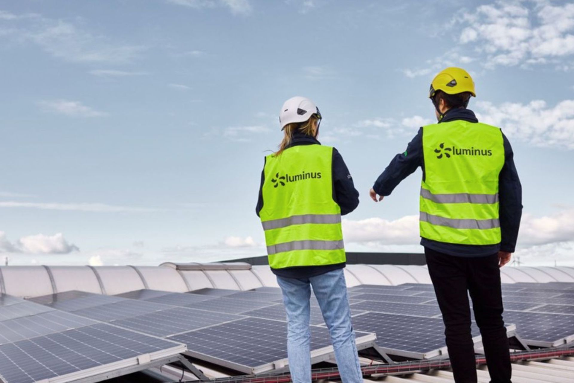 Deux techniciens portant des casques et des gilets de sécurité Luminus inspectent des panneaux solaires installés sur un toit, sous un ciel partiellement nuageux.