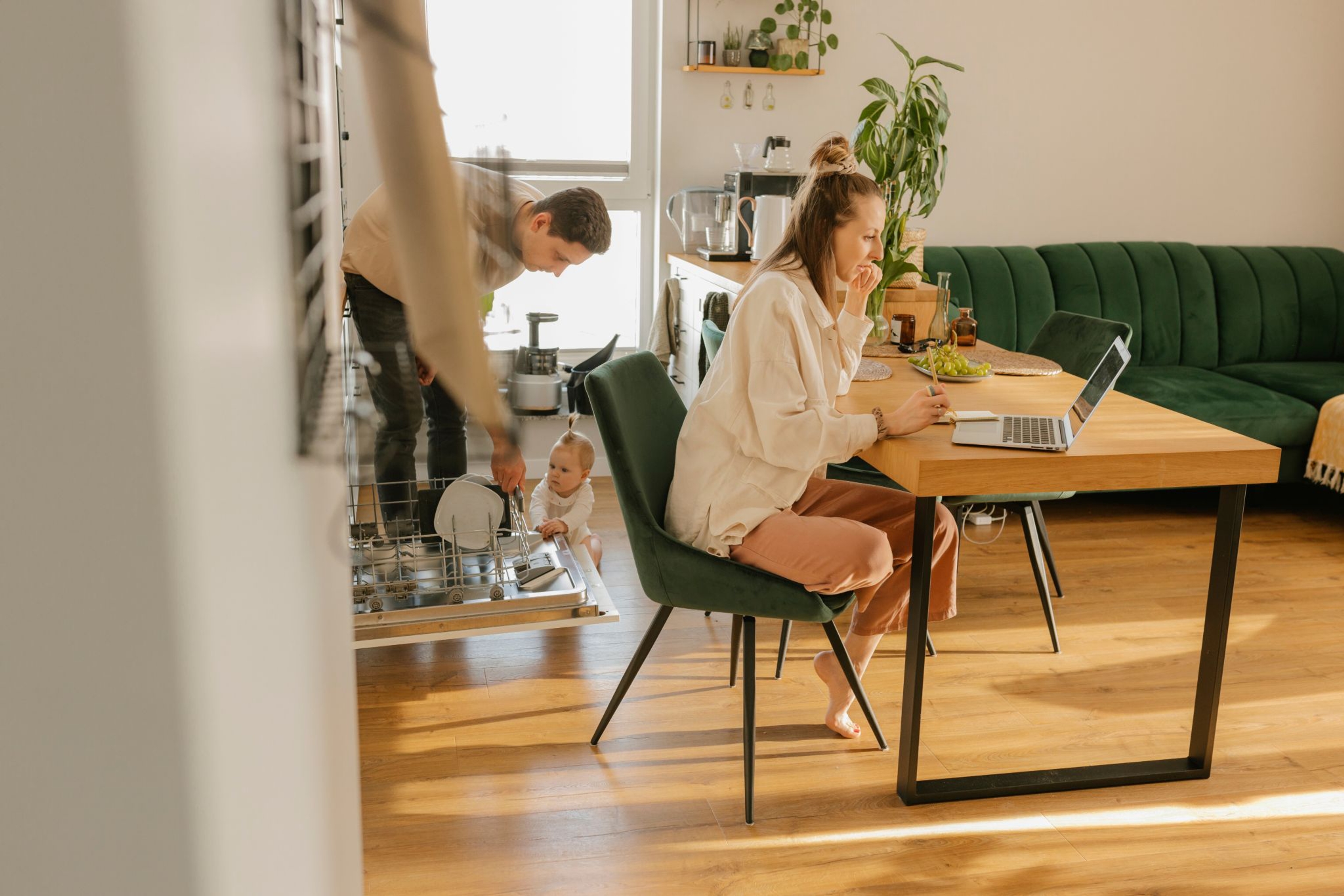 Une femme travaille sur un ordinateur portable à la table de la salle à manger. Un homme charge le lave-vaisselle tandis qu'un bébé joue à proximité.