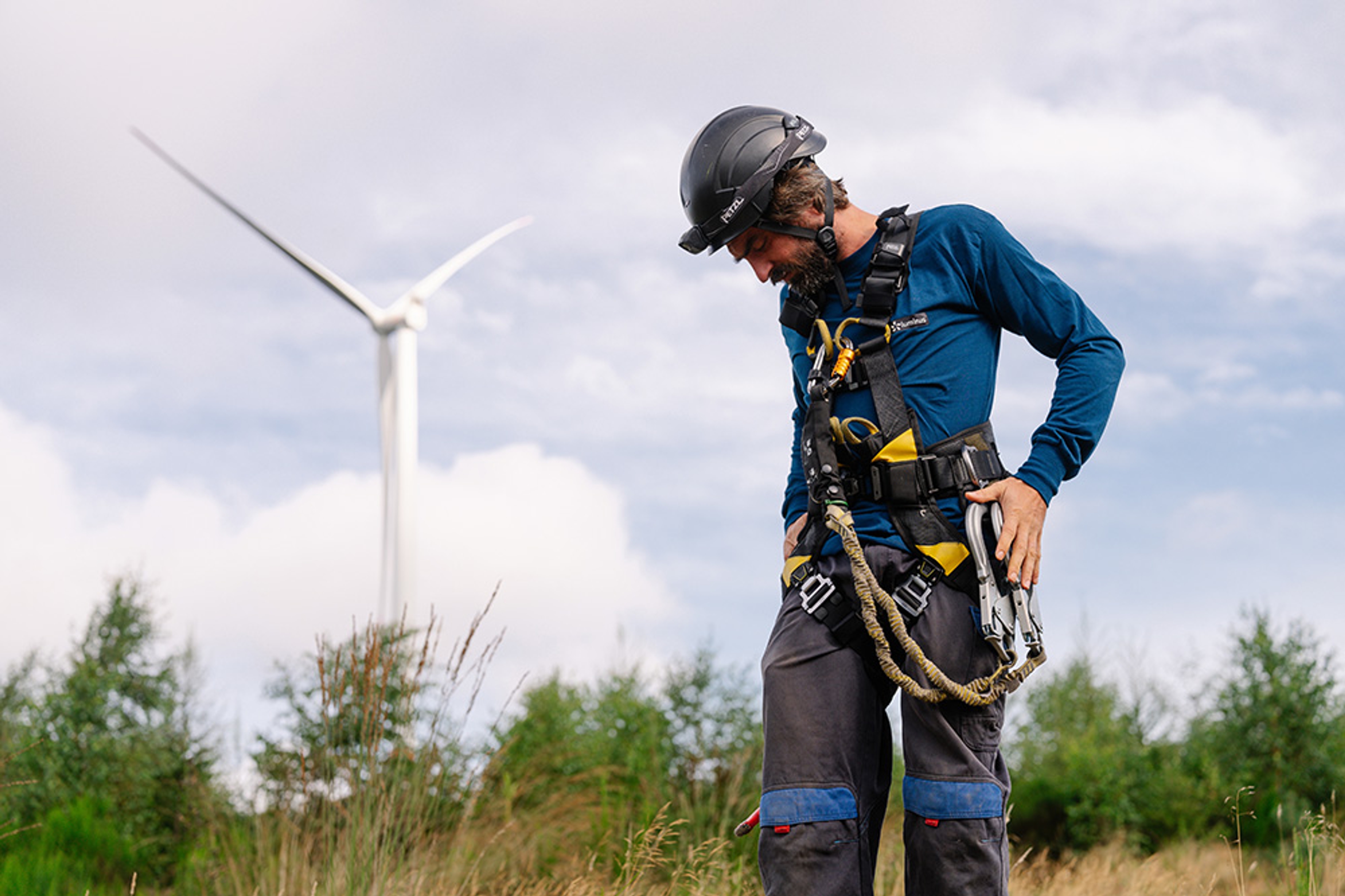 Windmolentechnieker met helm en klimuitrusting checkt zijn materiaal, windturbine in de achtergrond.