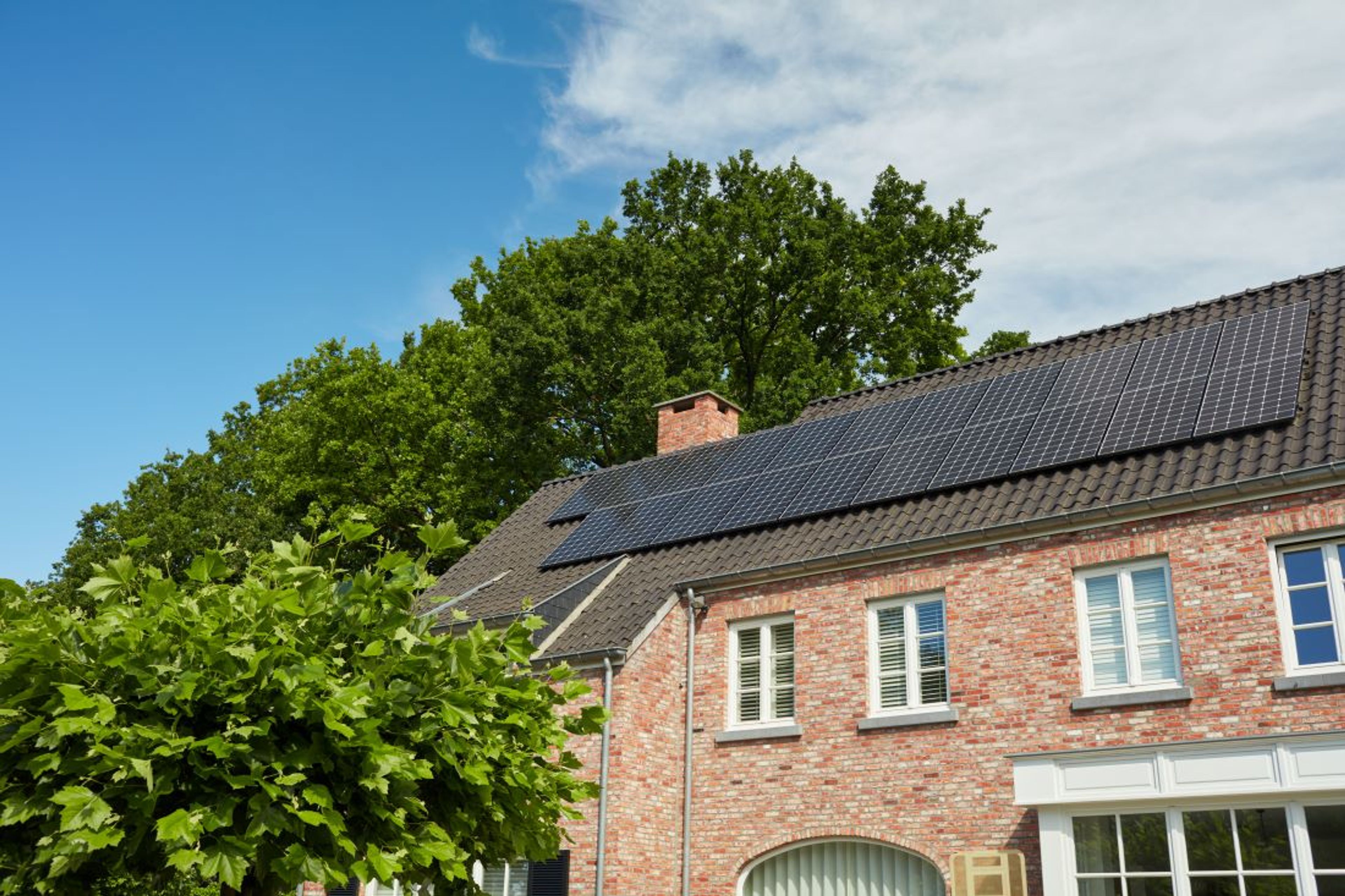 Maison en briques avec des panneaux solaires installés sur le toit incliné, entourée d’arbres verdoyants sous un ciel partiellement nuageux