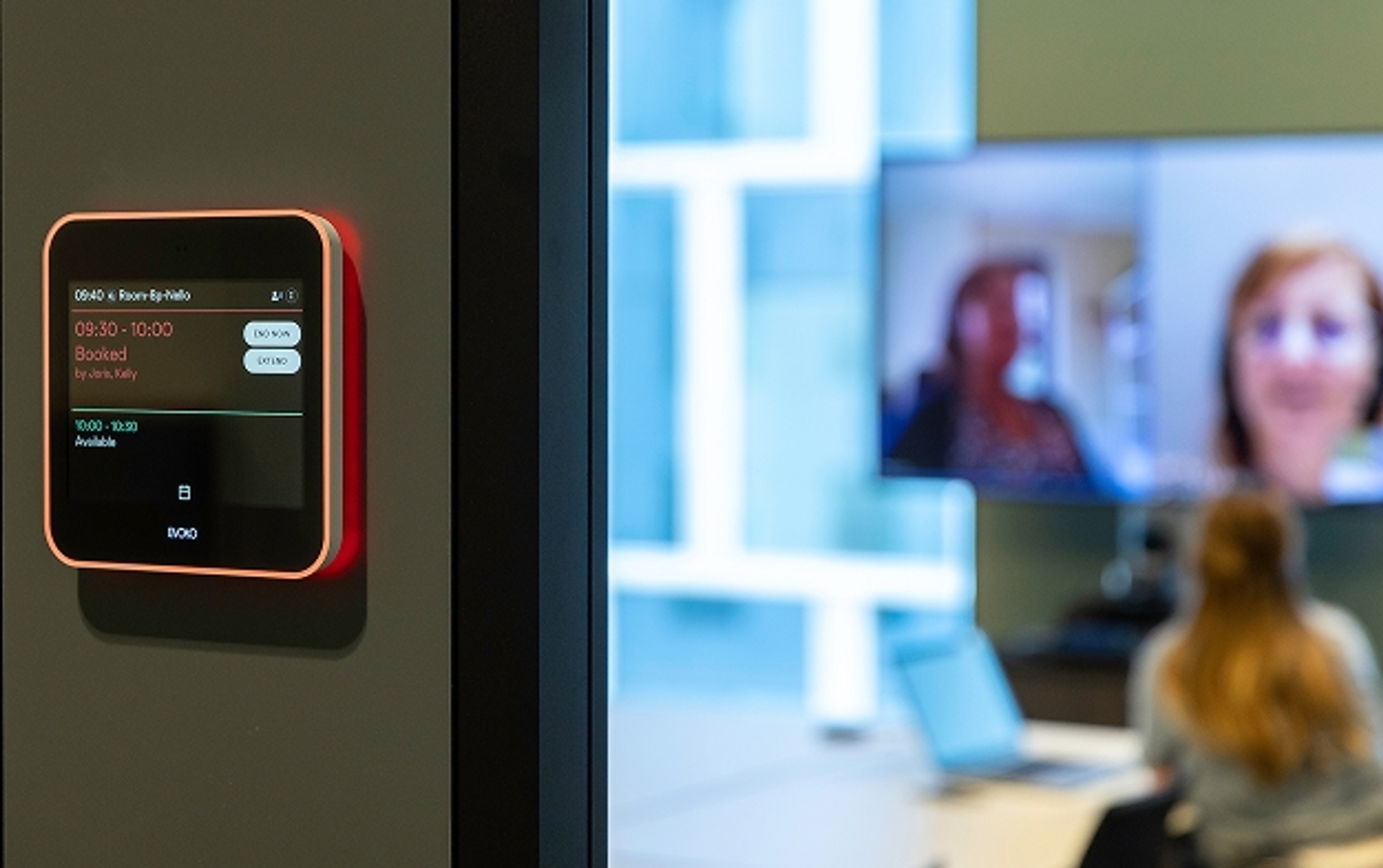 A digital meeting room schedule display shows a room is booked. In the background, a woman participates in a virtual meeting on a screen. Office atmosphere.