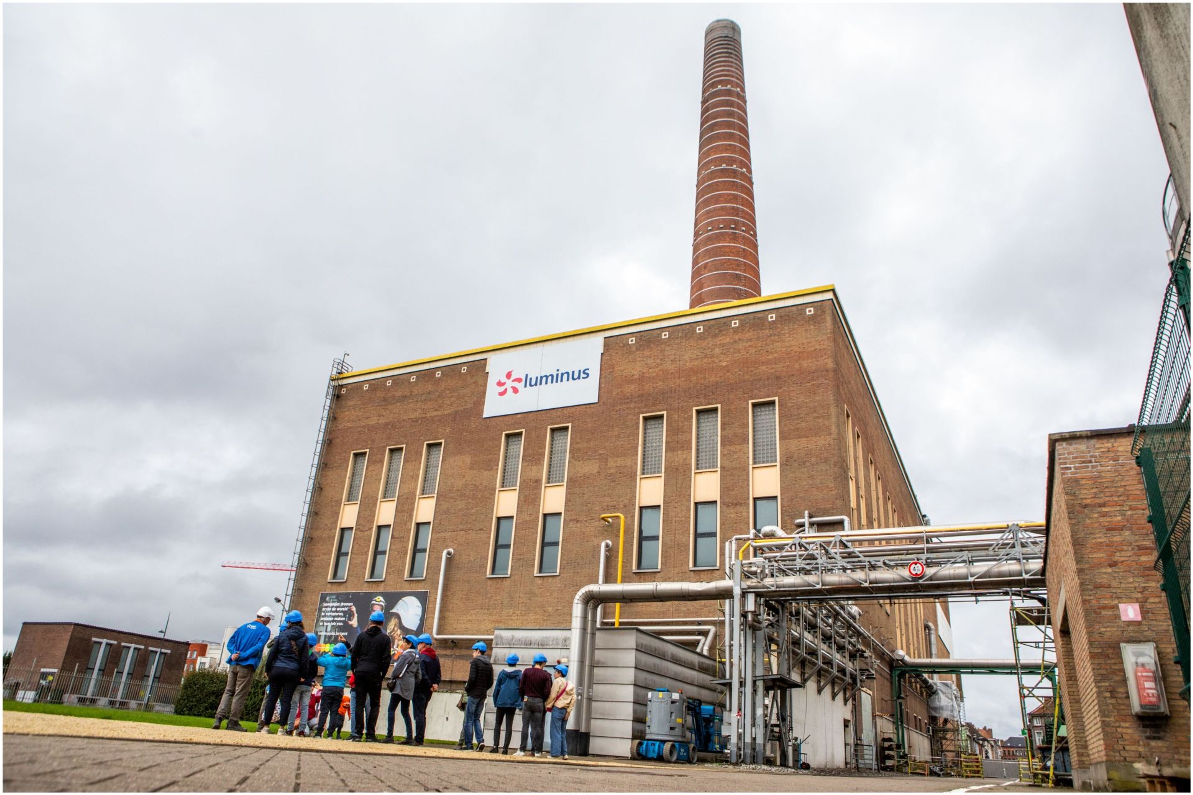 Centrale de Ham à Gand avec une haute cheminée en briques, des bâtiments techniques, un groupe à casques blues visitent le site.