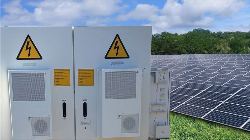 Links een batterij en rechts een veld vol zonnepanelen, groen gras, groene bomen en blauwe lucht.