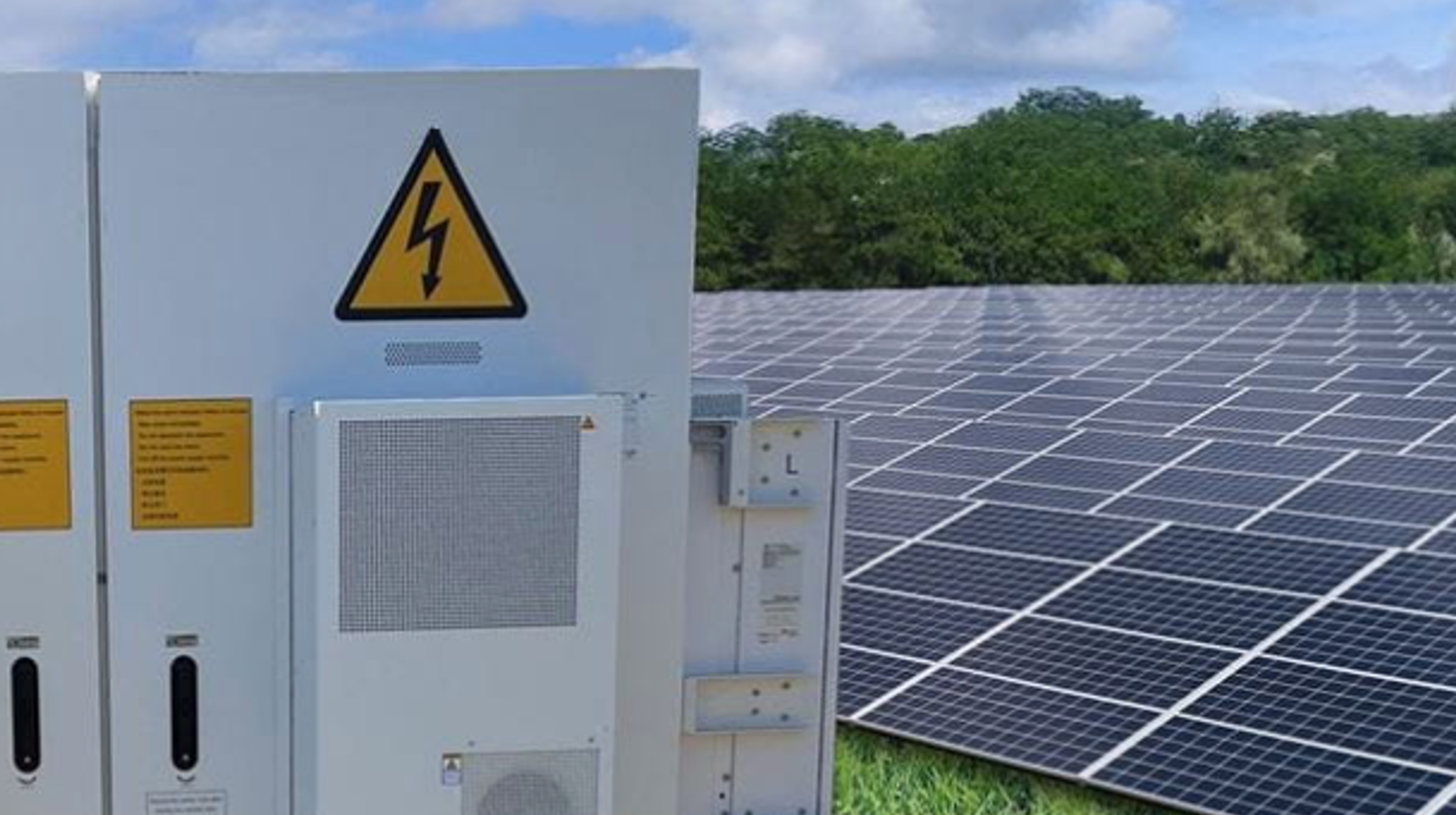 Links een batterij en rechts een veld vol zonnepanelen, groen gras, groene bomen en blauwe lucht.