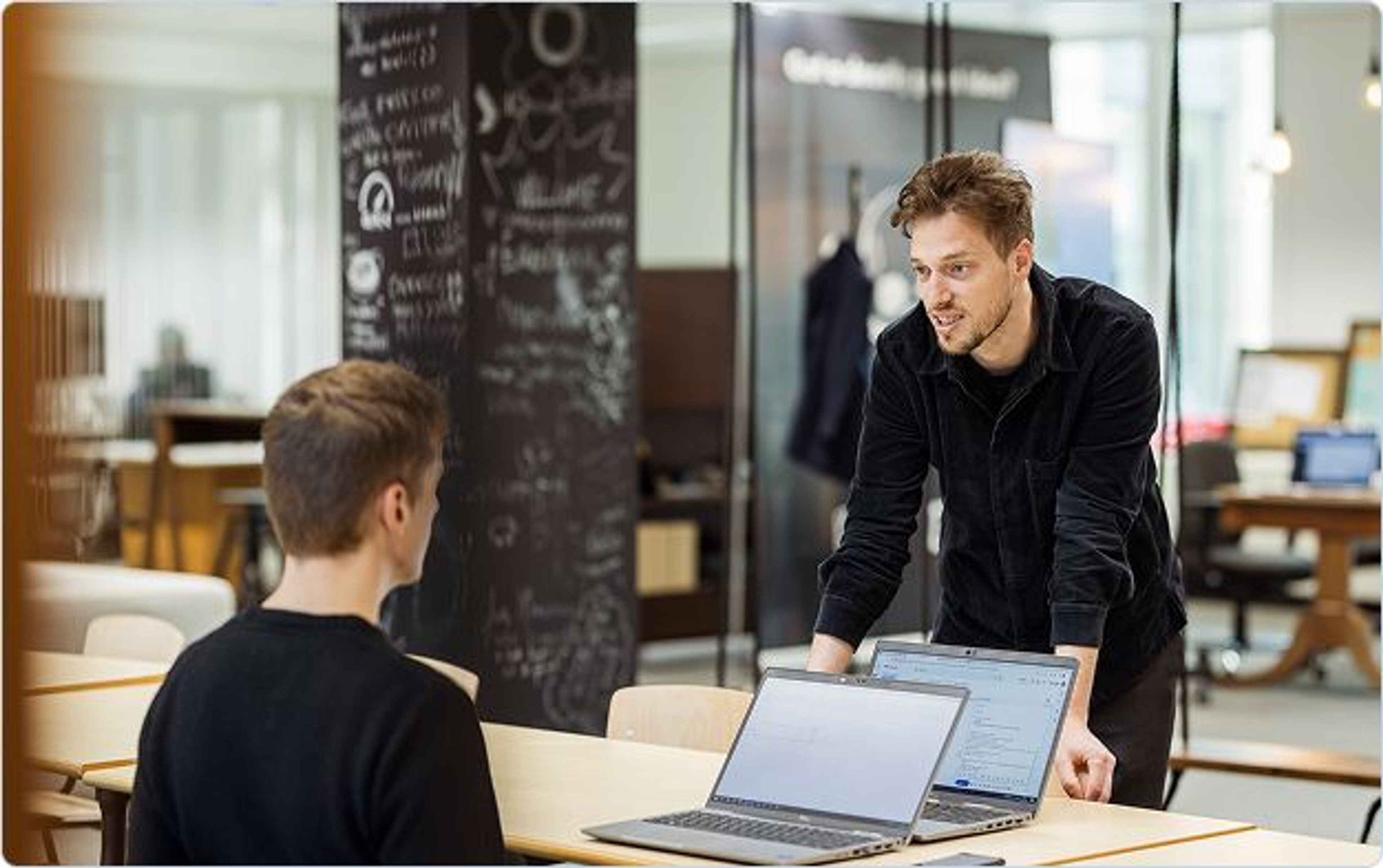 2 young men are having a chat in the office, one seated with 2 laptops, one standing