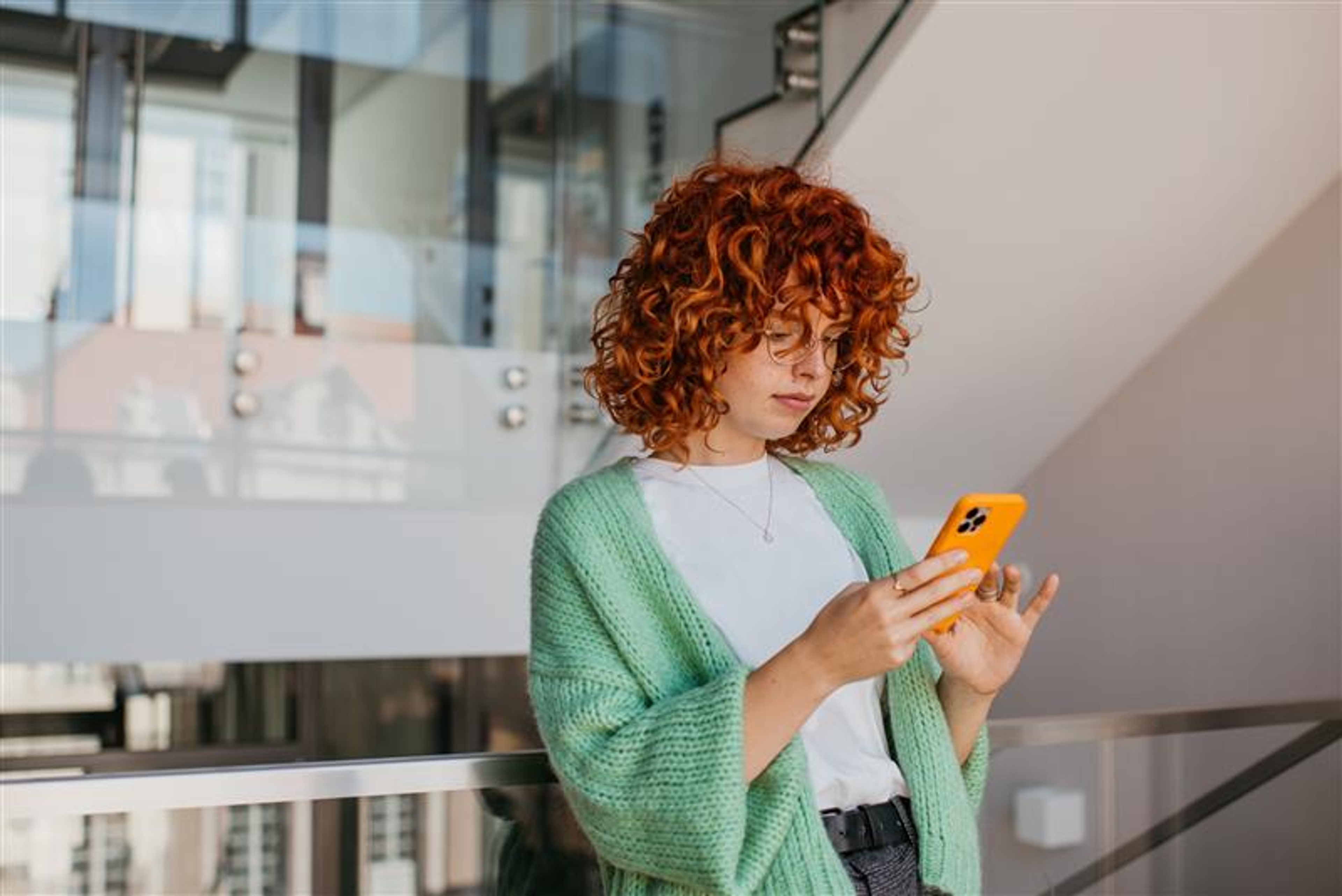 Jonge vrouw met oranje krullen en groene trui checkt haar smartphone vlakbij een trap met glazen leuning.