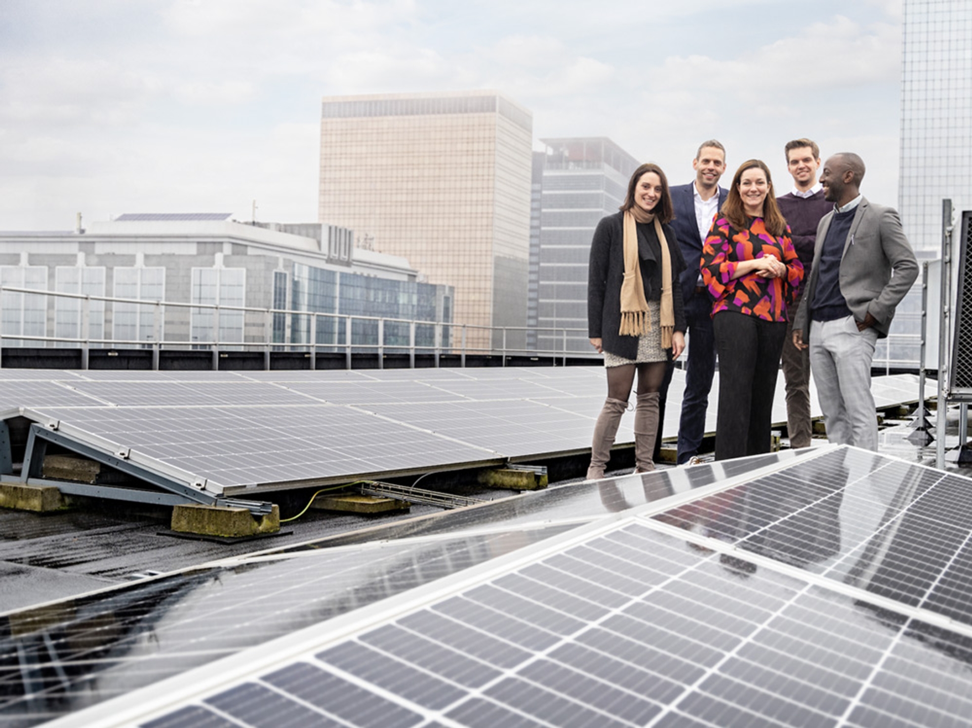 Groupe de cinq personnes souriantes en tenue professionnelle, debout sur un toit équipé de panneaux solaires, avec des immeubles de bureaux en arrière-plan
