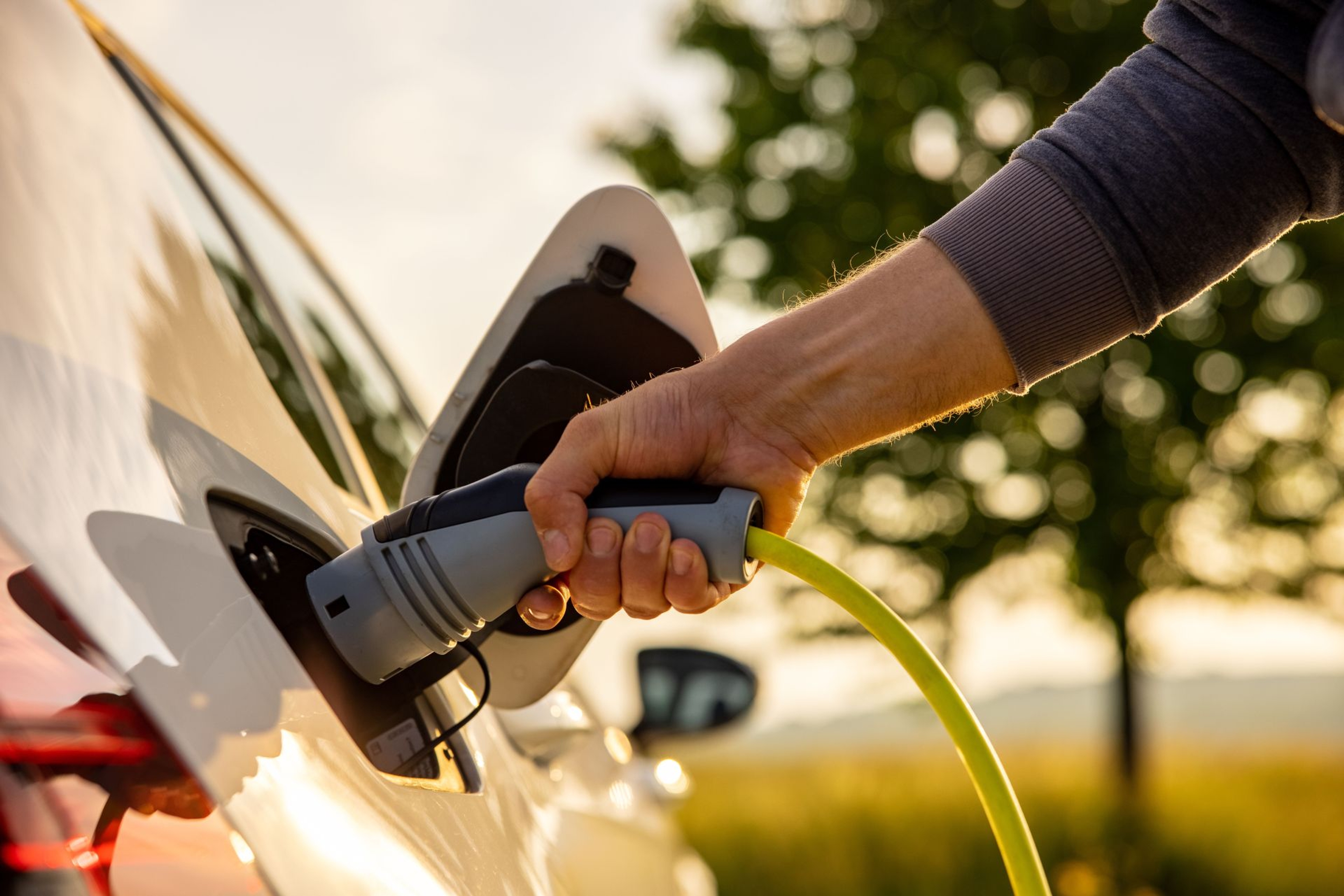 Zonnig licht, we zien een mannenhand die de stekker in een e-wagen steekt.