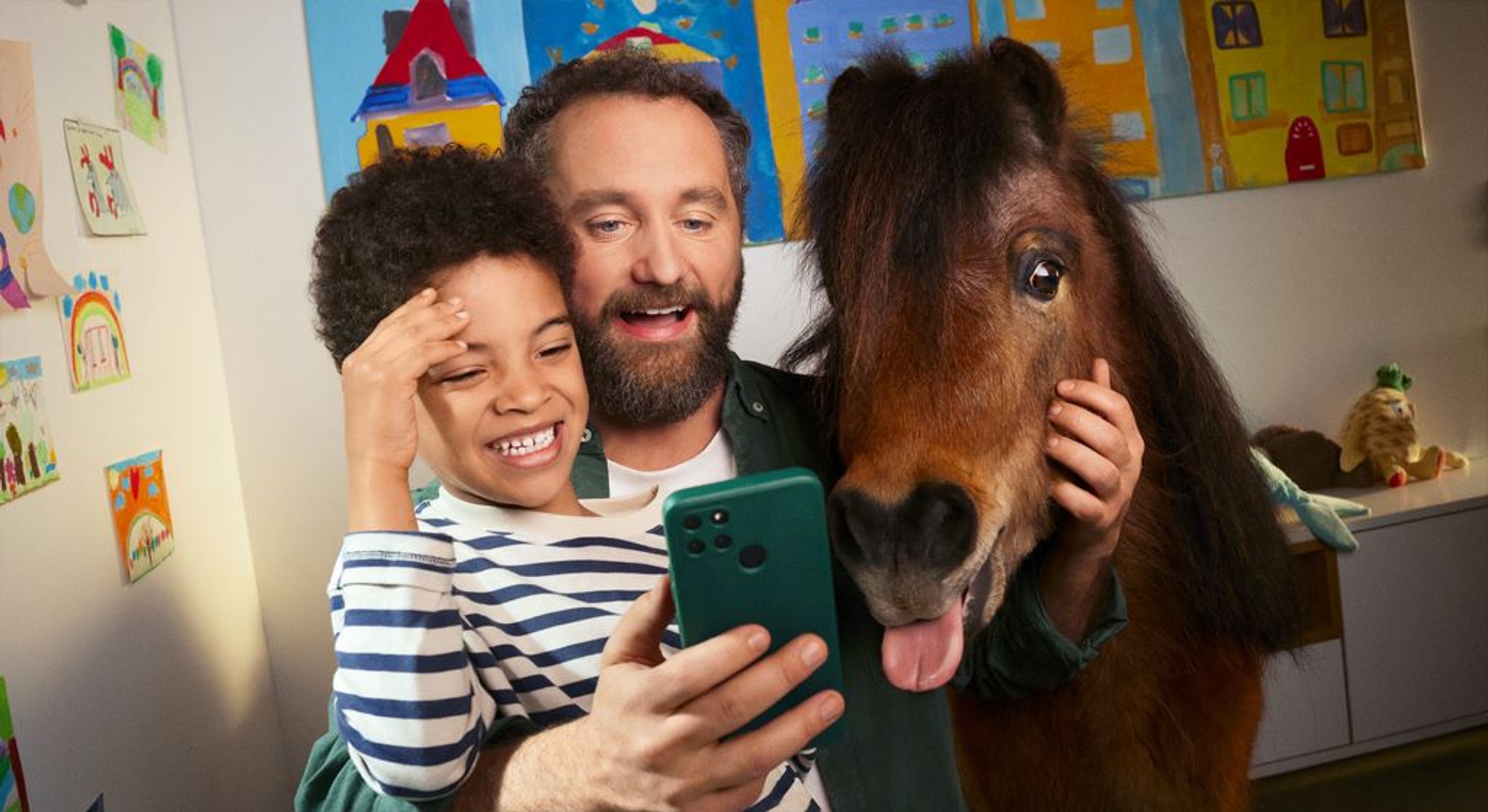 Man met baard, jongetje en pony in woonkamer, man kijkt op smartphone.