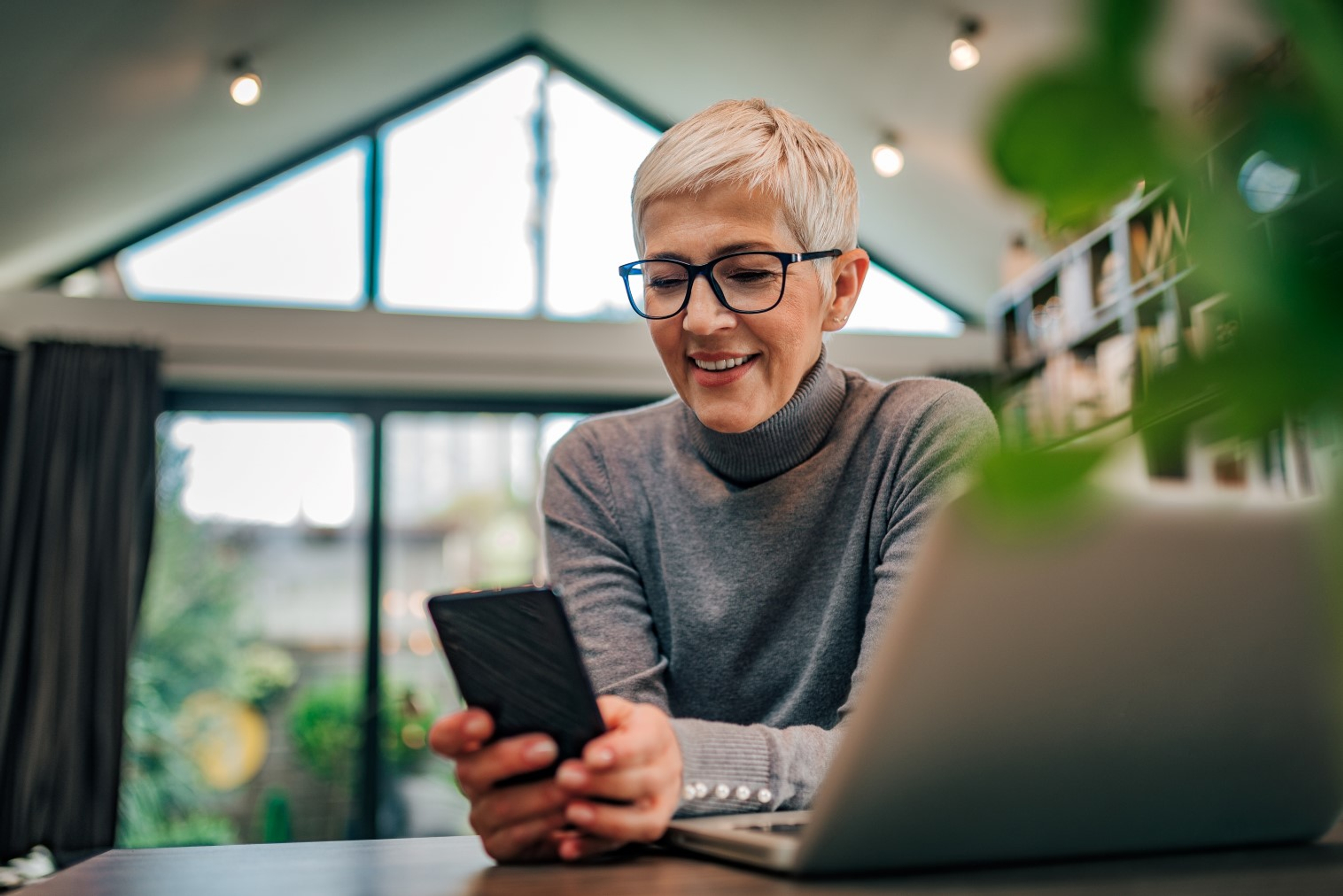 Dame met kort wit haar en zwarte bril in haar moderne woonkamer, kijkt op smartphone bij laptop.