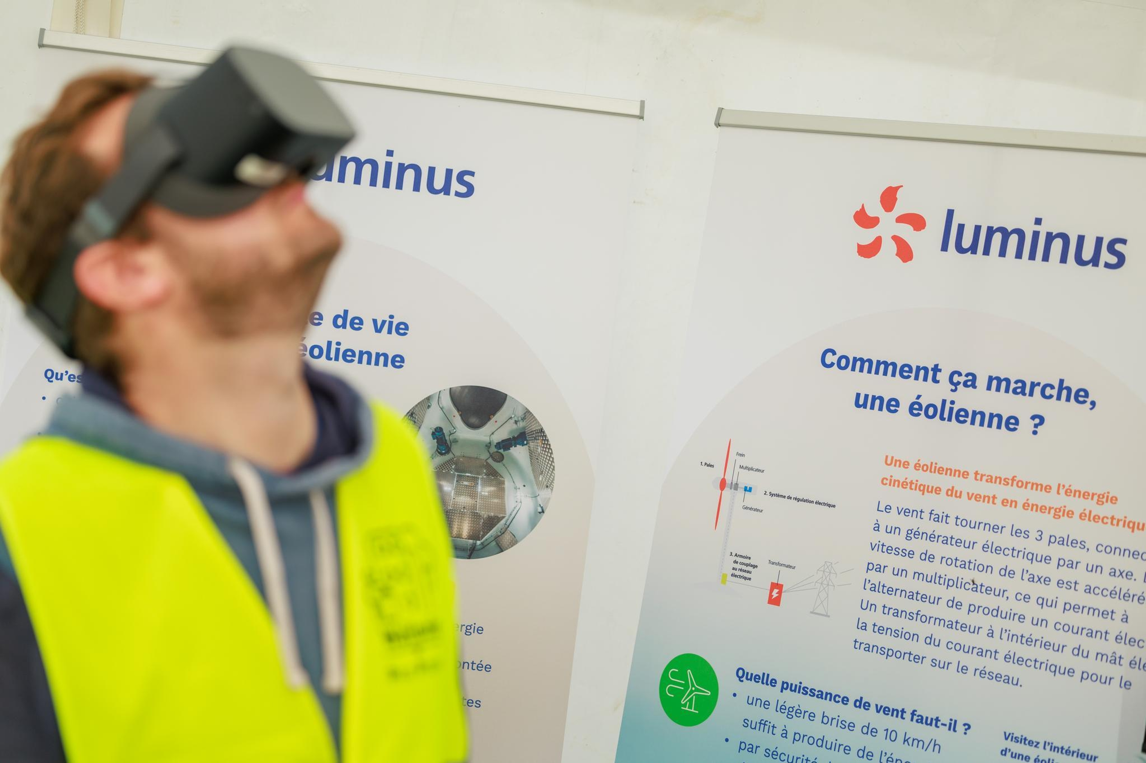 Een persoon met een VR-headset en een geel veiligheidsvest kijkt omhoog. Achter hen hangen twee informatieposters over windturbines, met diagrammen en het logo ‘Luminus’. De scène straalt een gevoel van leren en innovatie uit.