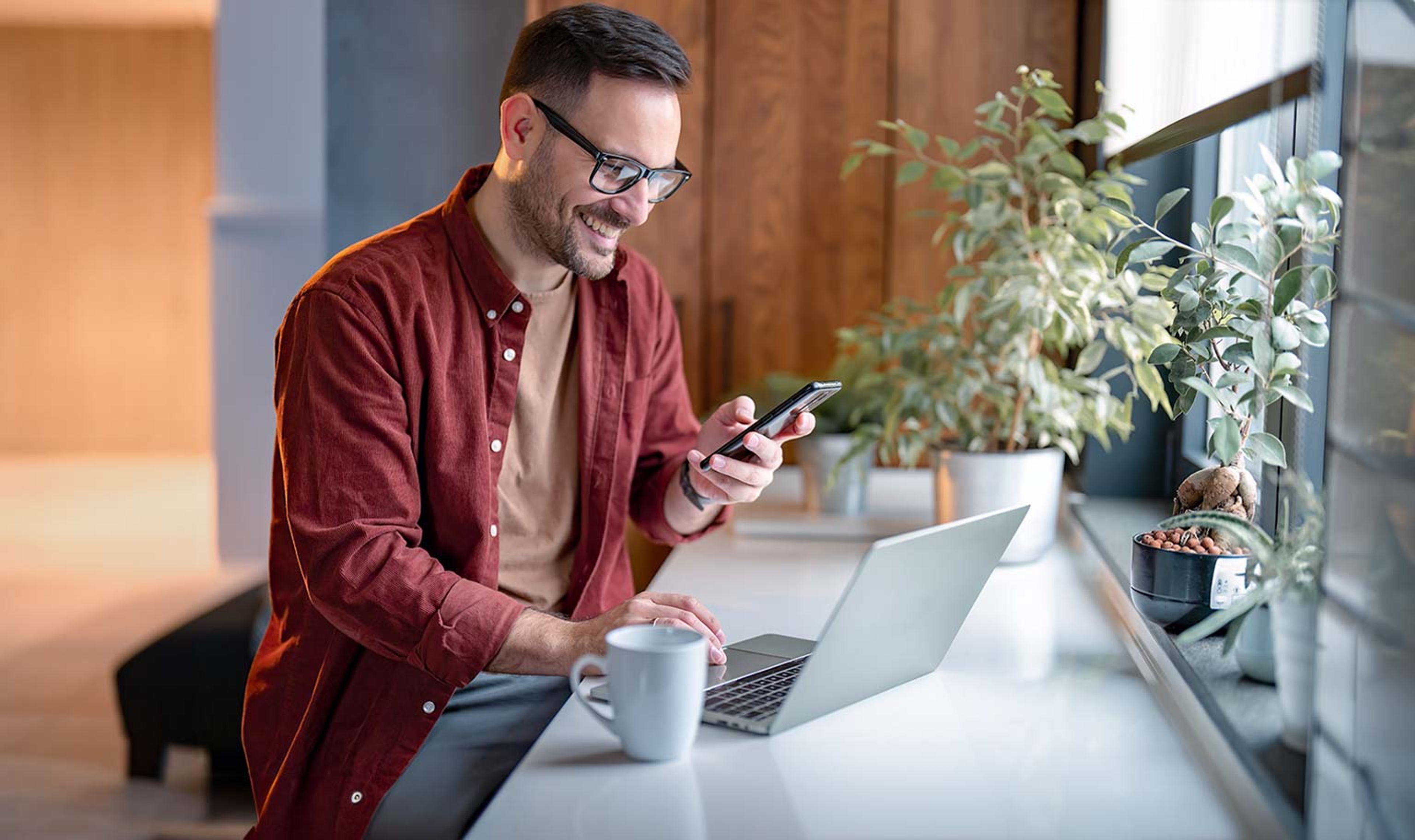 Man in een rood hemd kijkt lachend op zijn smartphone, één hand aan de laptop.