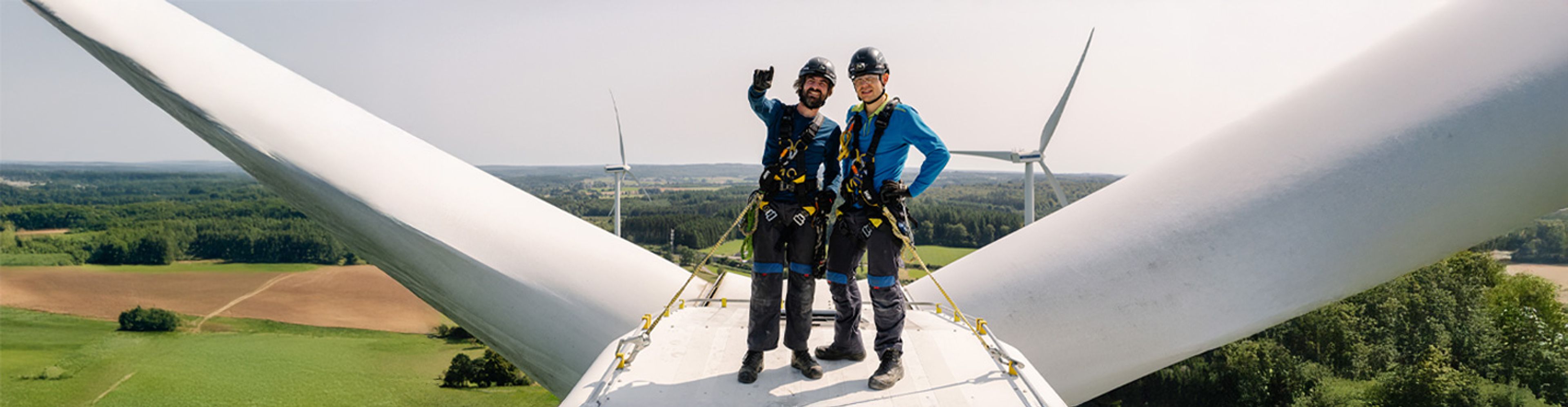 Twee werknemers in veiligheidskleding staan bovenop een windturbine, glimlachend en zwaaiend. Op de achtergrond zijn uitgestrekte groene velden en nog meer turbines te zien onder een heldere hemel.