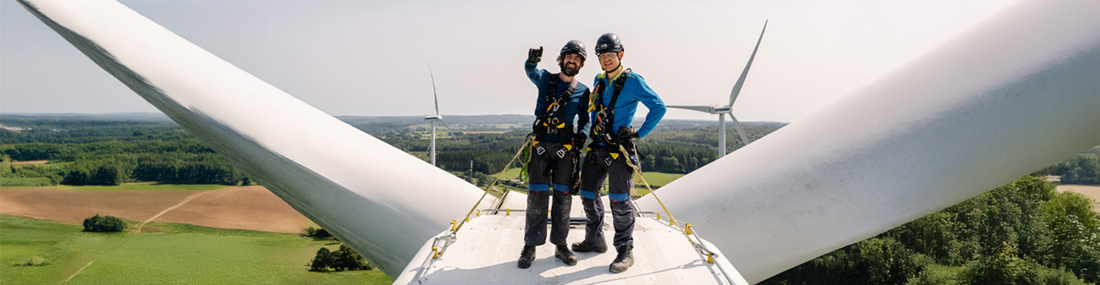 Twee werknemers in veiligheidskleding staan bovenop een windturbine, glimlachend en zwaaiend. Op de achtergrond zijn uitgestrekte groene velden en nog meer turbines te zien onder een heldere hemel.
