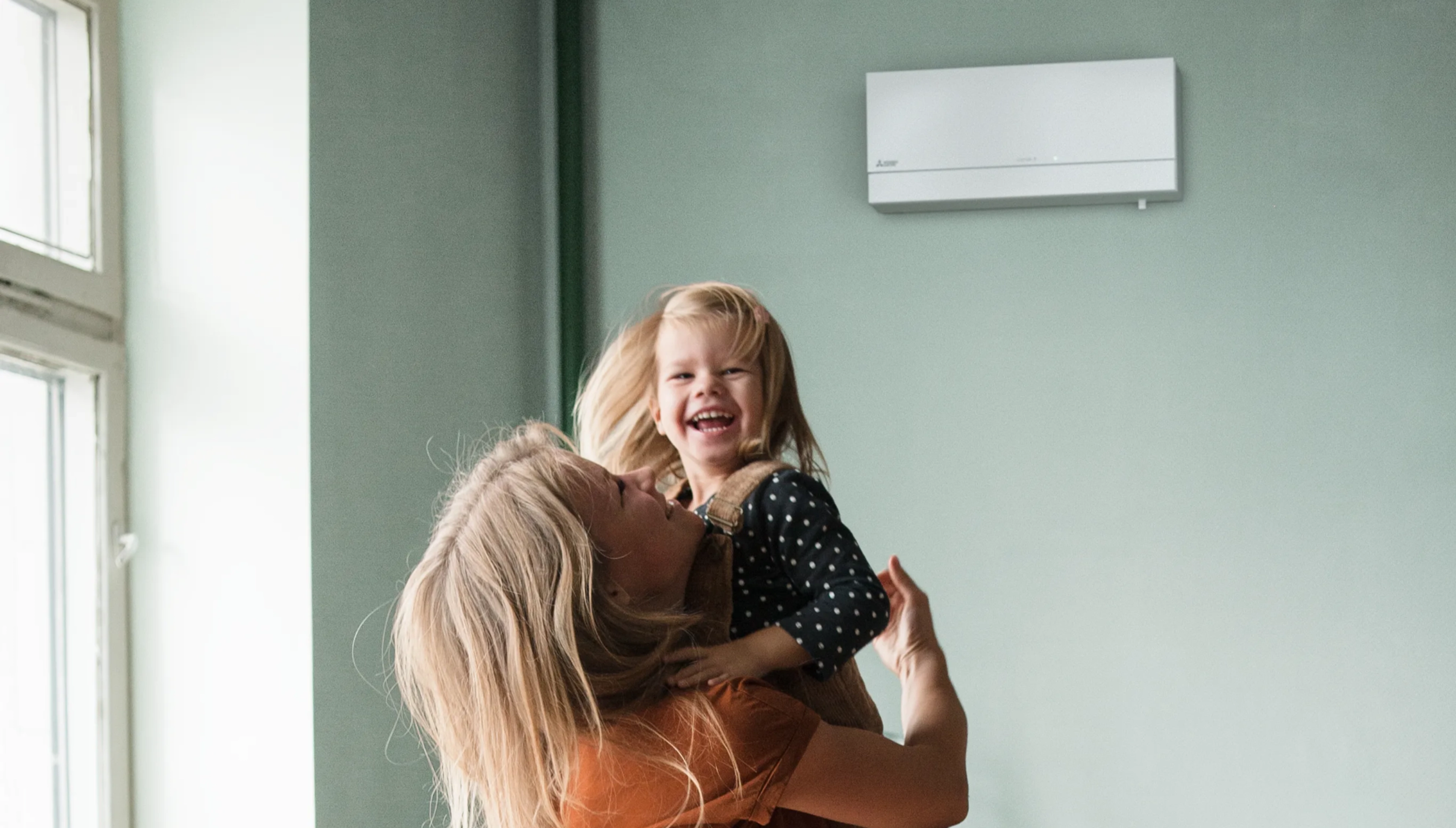 Jonge vrouw tilt klein meisje op, ze lachen. Ze bevinden zich in een living met lichtrgoen geschilderde muren en een binnenunit van een lucht-lucht warmtepomp.