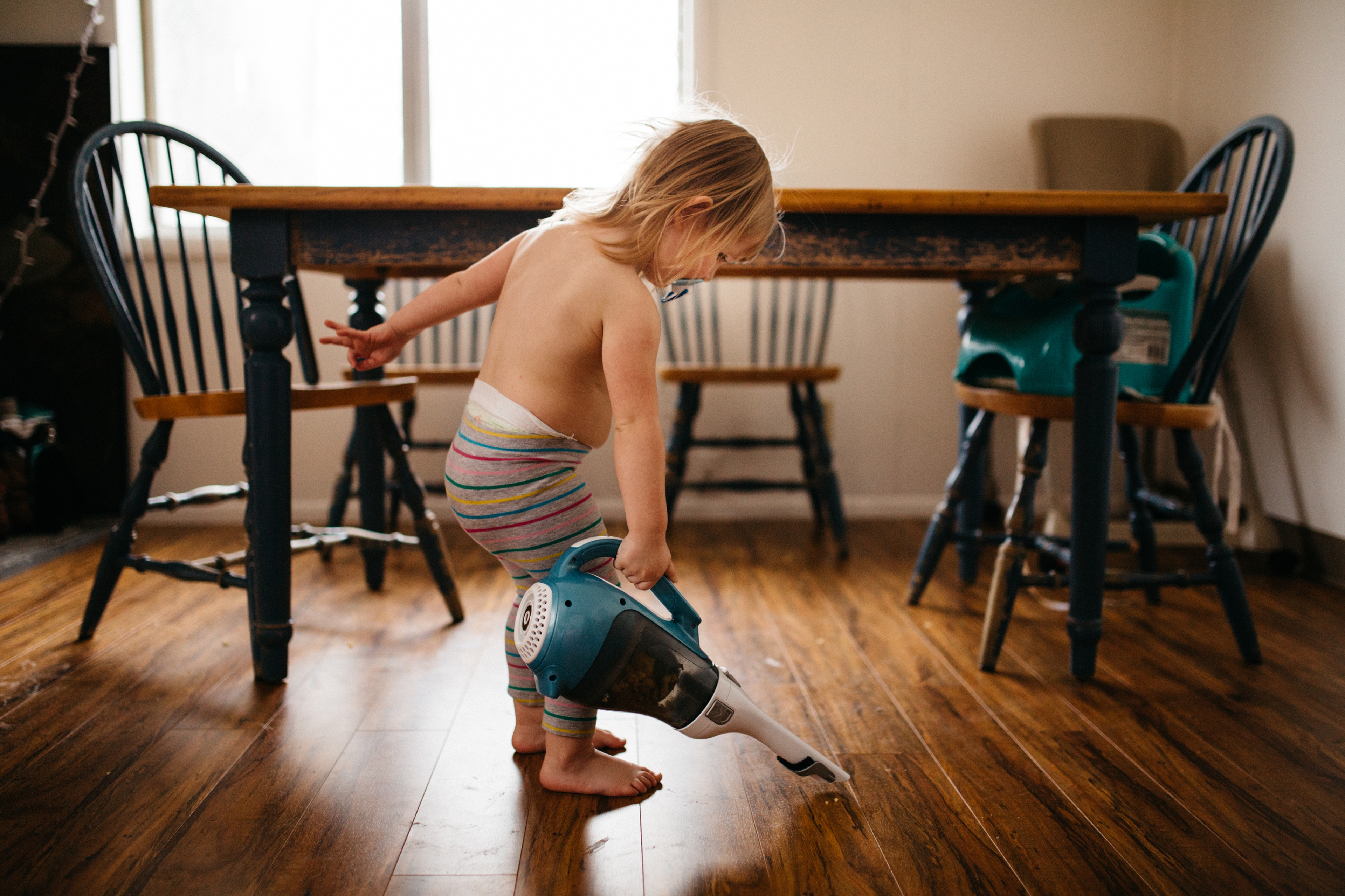 Un jeune enfant passe l'aspirateur sur le sol d'une pièce de vie, près d'une table et de chaises.