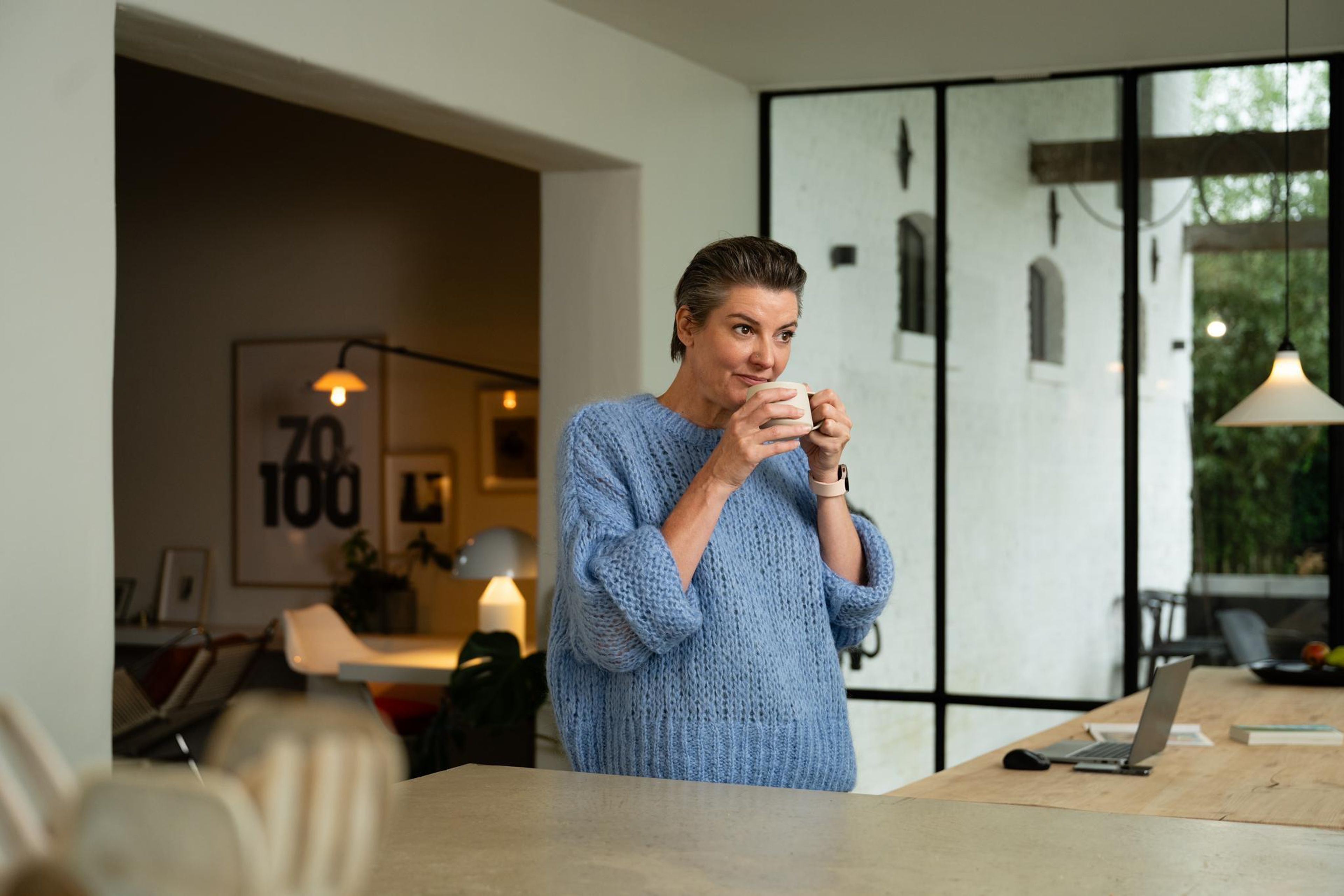 Une femme vêtue d'un pull bleu confortable savoure une tasse de café dans une cuisine moderne. La lumière naturelle qui pénètre par les grandes fenêtres crée une atmosphère sereine.