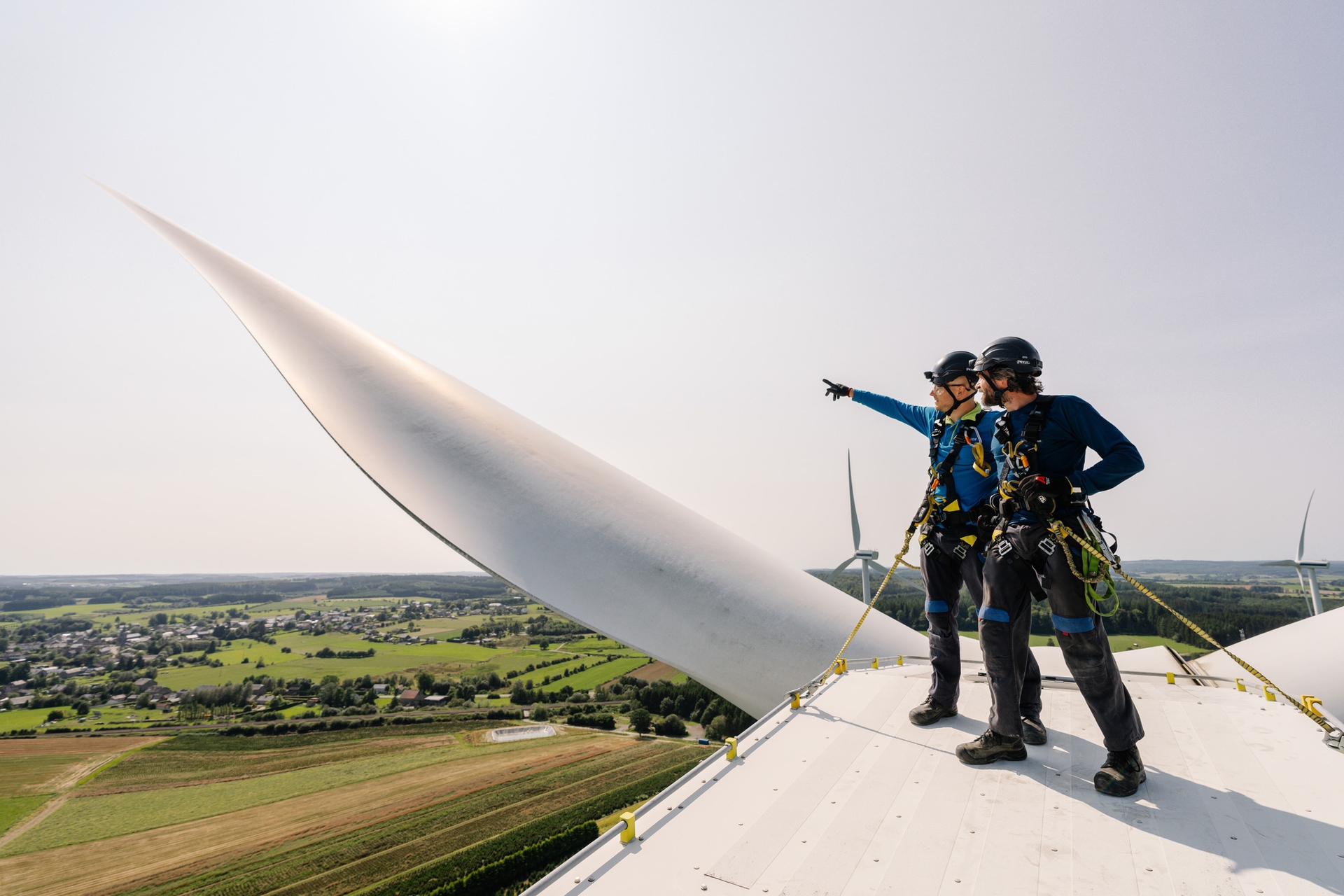 Twee werknemers in veiligheidskleding staan bovenop een windturbine, waarvan er één naar het landschap beneden wijst. De scène straalt teamwork en innovatie uit.