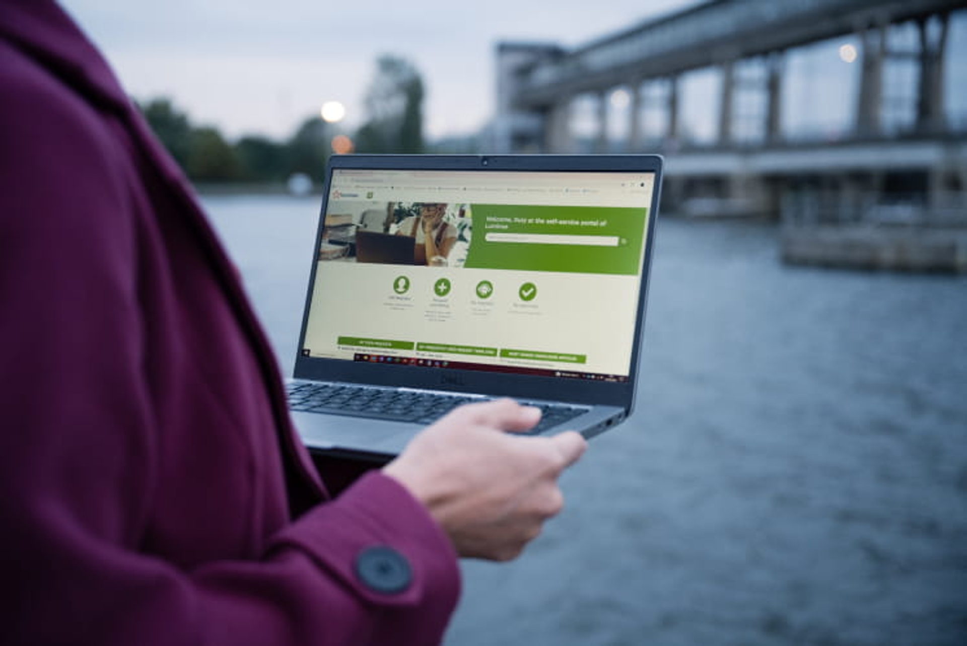 woman with purple coat holds a laptop, in the back we see a waterpower installation on the river.
