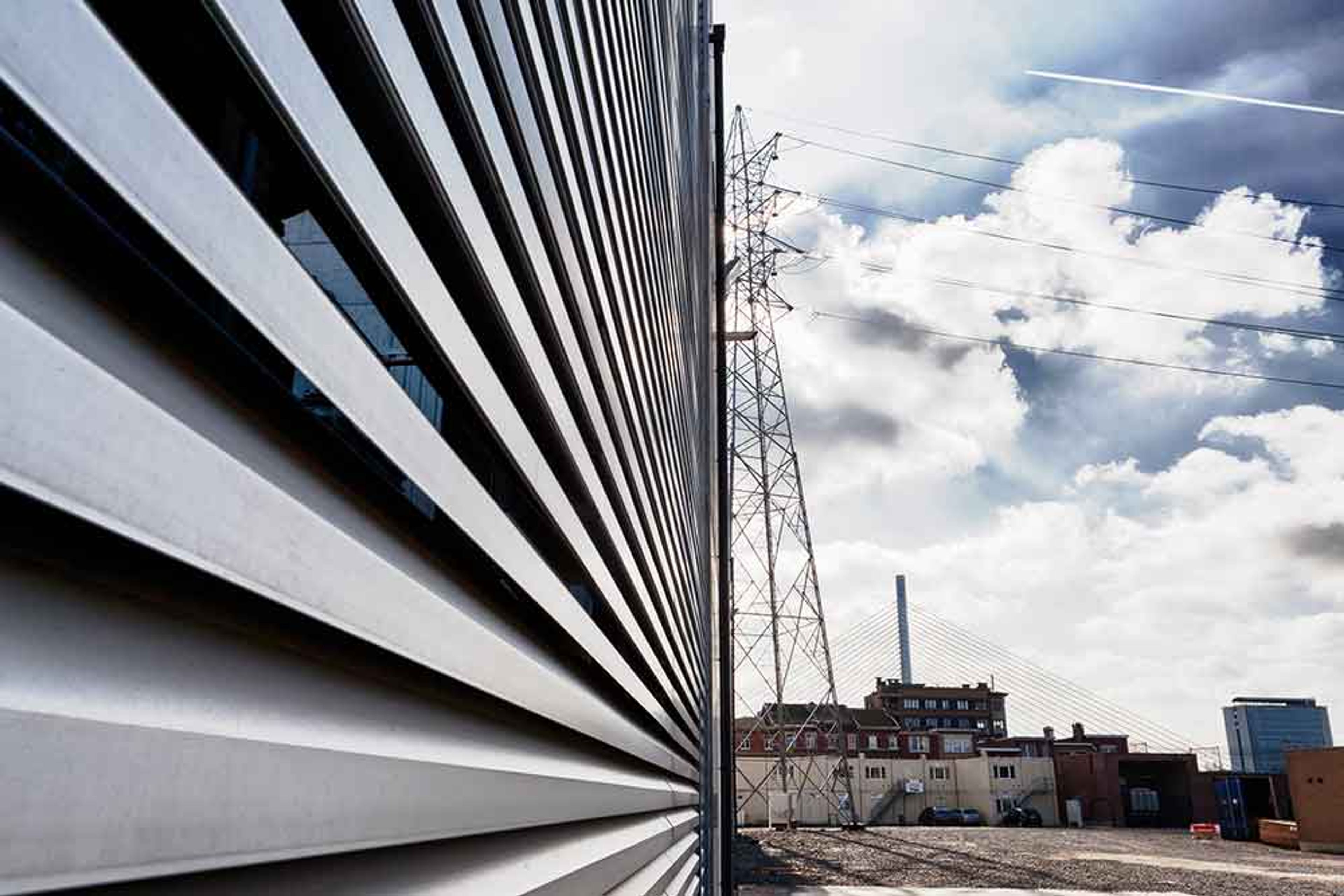 Vue d’un bâtiment industriel à façade métallique nervurée, avec en arrière-plan un pylône électrique, des câbles haute tension, des bâtiments urbains et un ciel partiellement nuageux