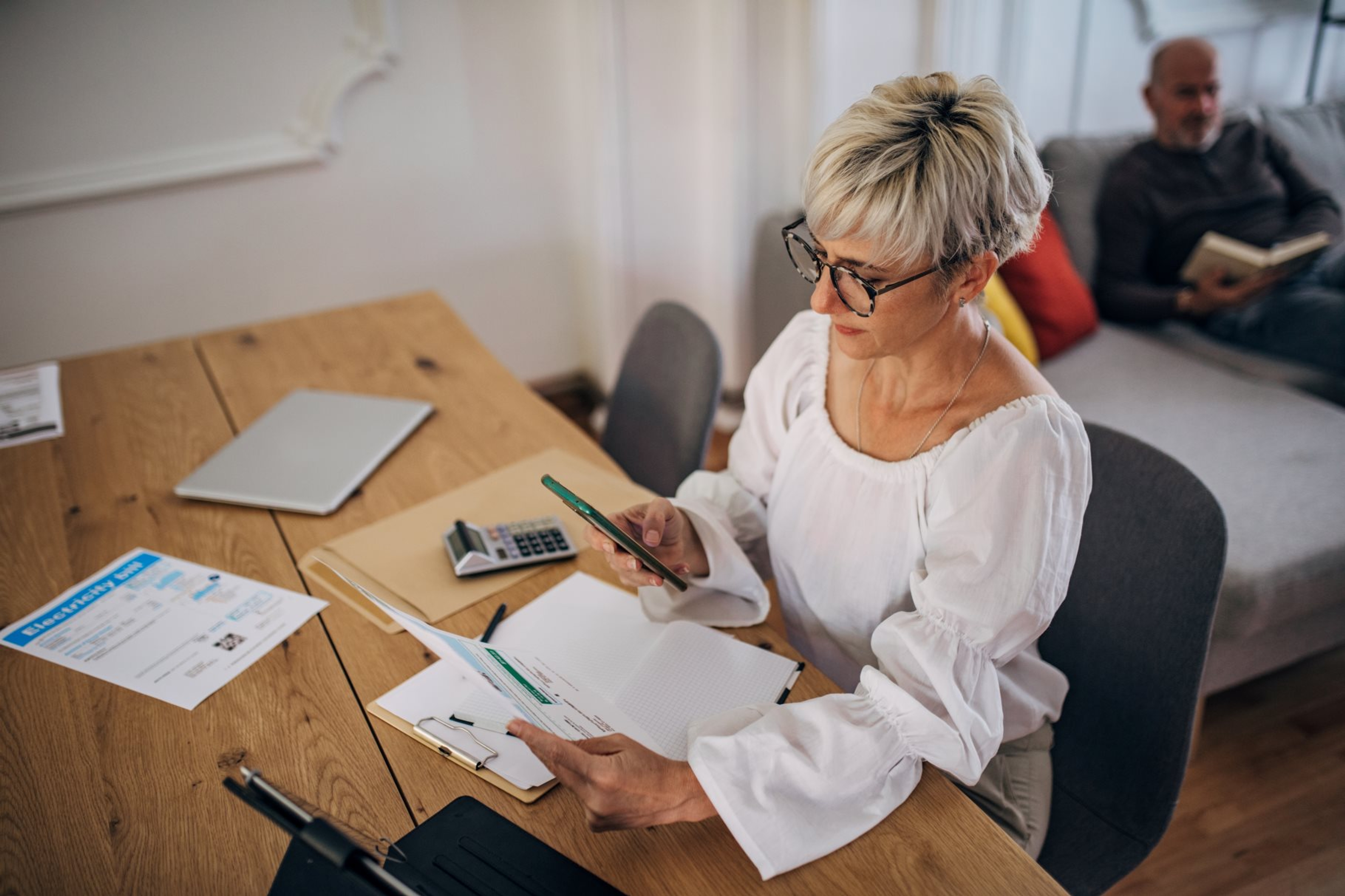 Dame met witte bloes en kort blond haar bekijkt papier en heeft smartphone in de andere hand, laptop op tafel.