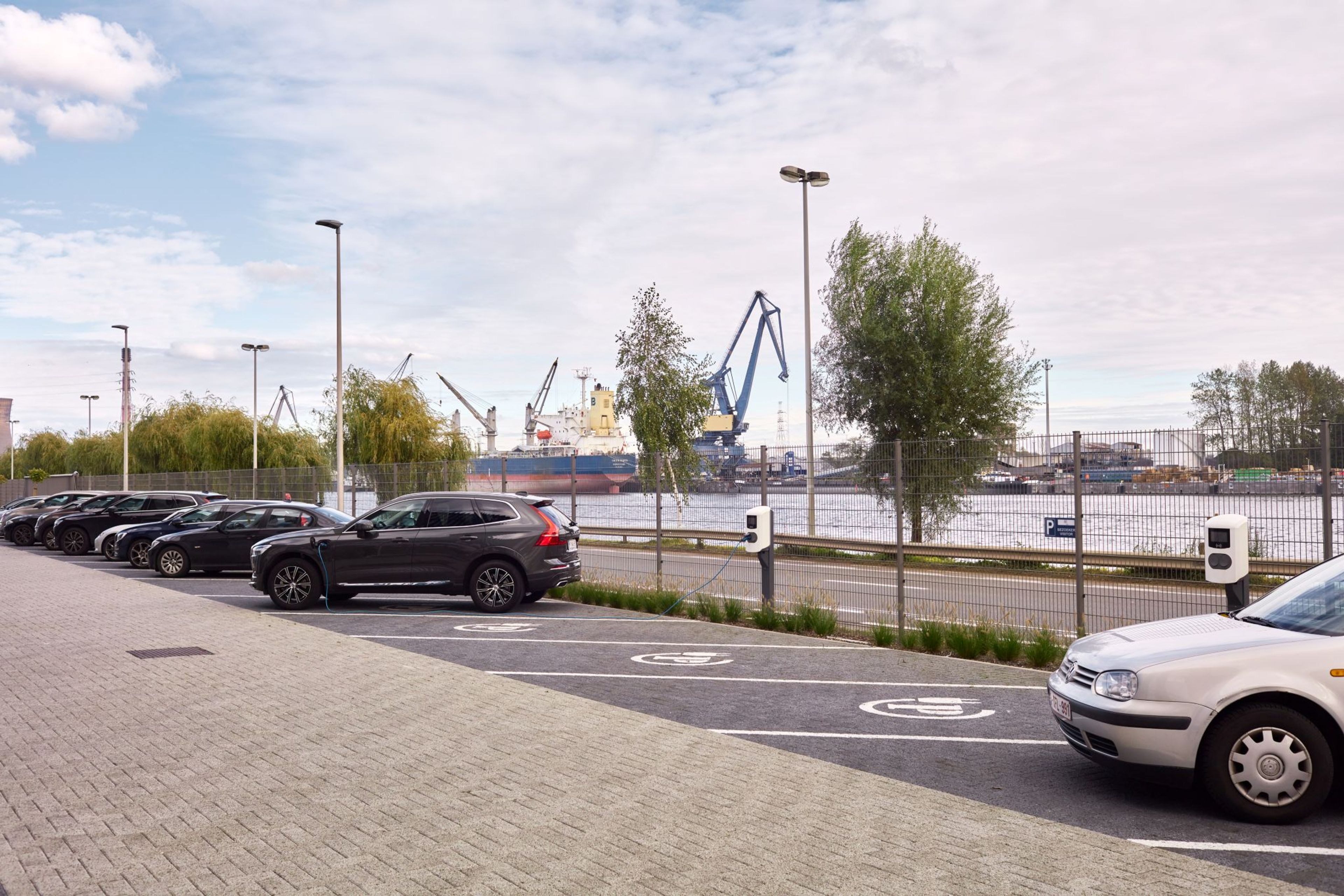 Parking d'entreprise équipé de bornes de recharge pour véhicules électriques, avec vue sur un port industriel en arrière-plan