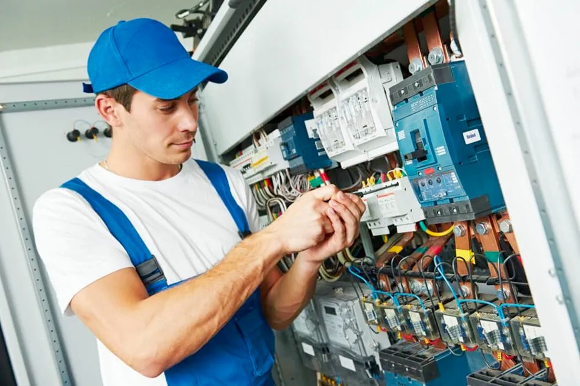 Électricien en uniforme bleu effectuant des réglages sur un tableau électrique rempli de câblages et de disjoncteurs